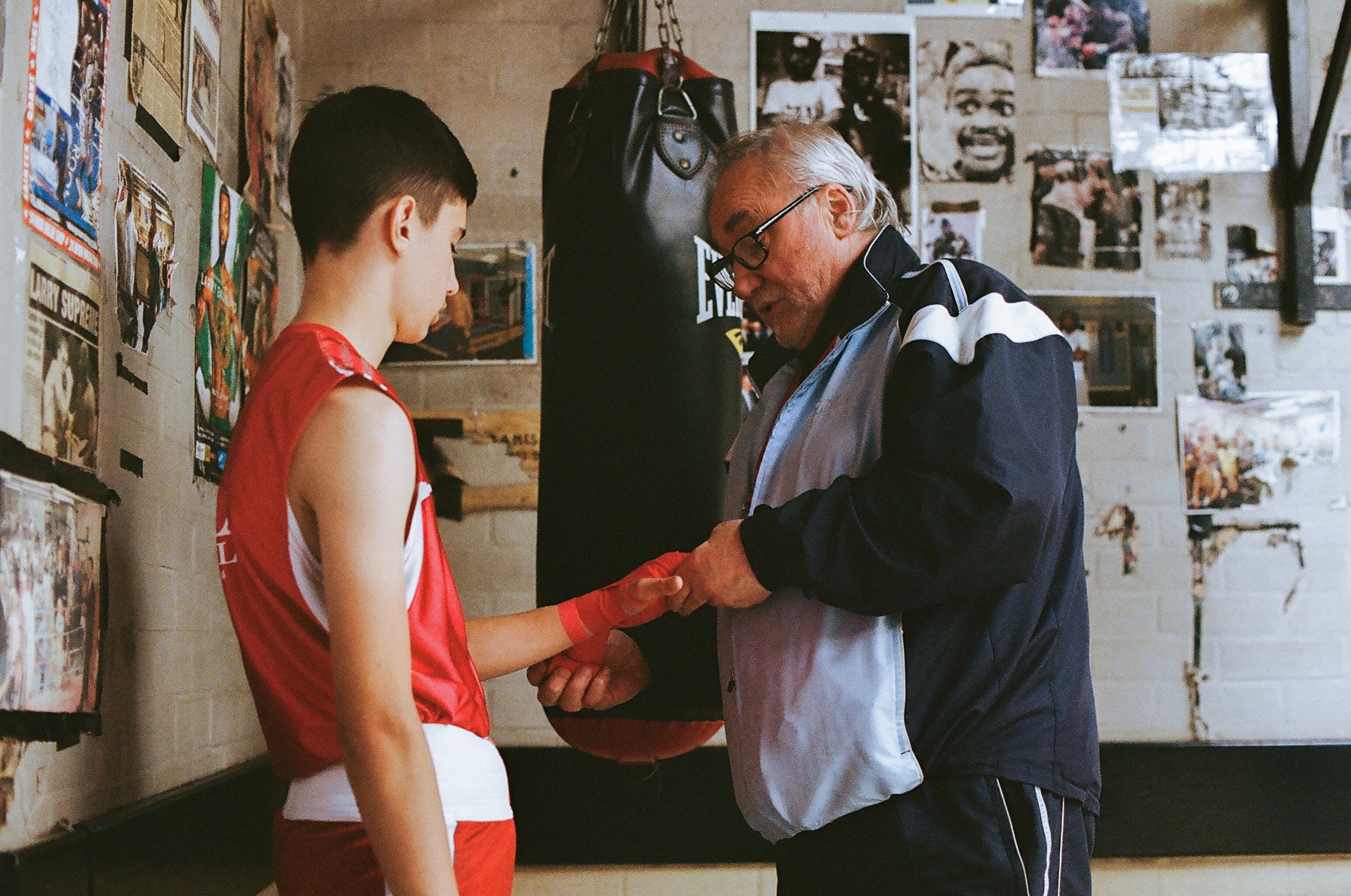 Older man wrapping hands of young boxer in red vest inside gym with photographs and posters covering walls.