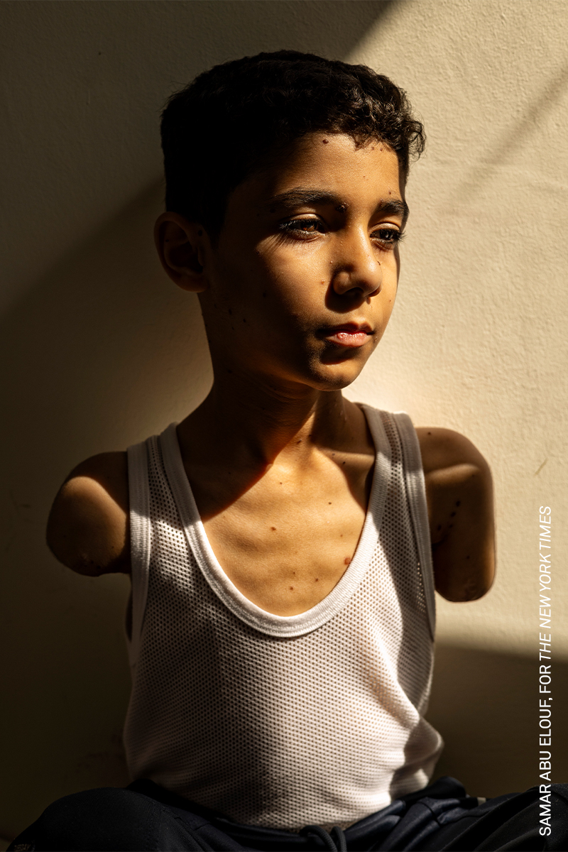 Young person with dark features gazing intently, wearing a white vest top against a plain background.