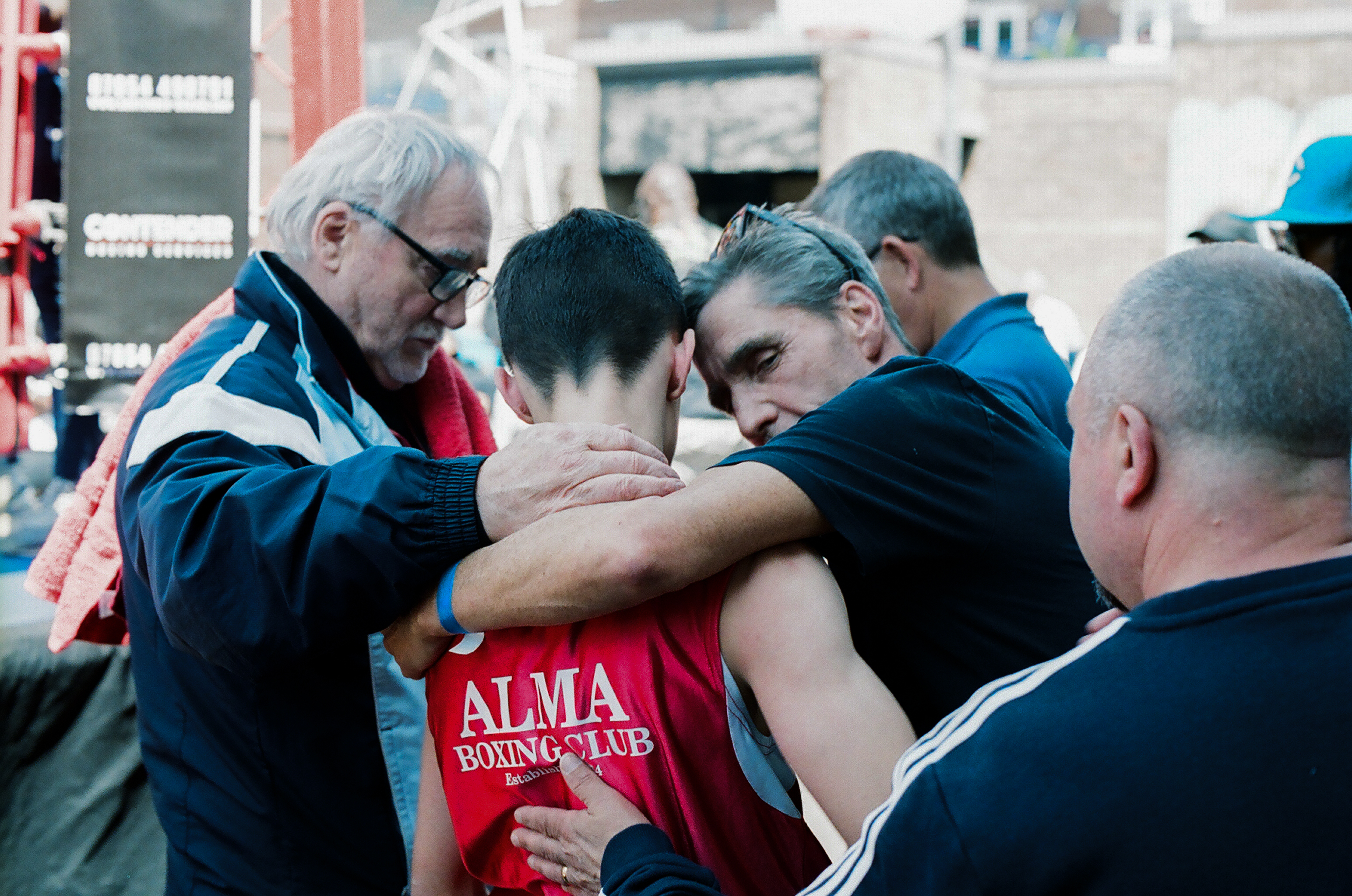 Boxing trainers and corner team huddle around fighter in red vest, providing support and instruction between rounds.