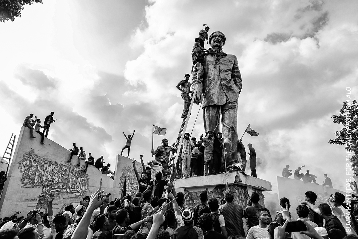 Monumental statue of a group figure amidst a crowd of onlookers against a cloudy sky.