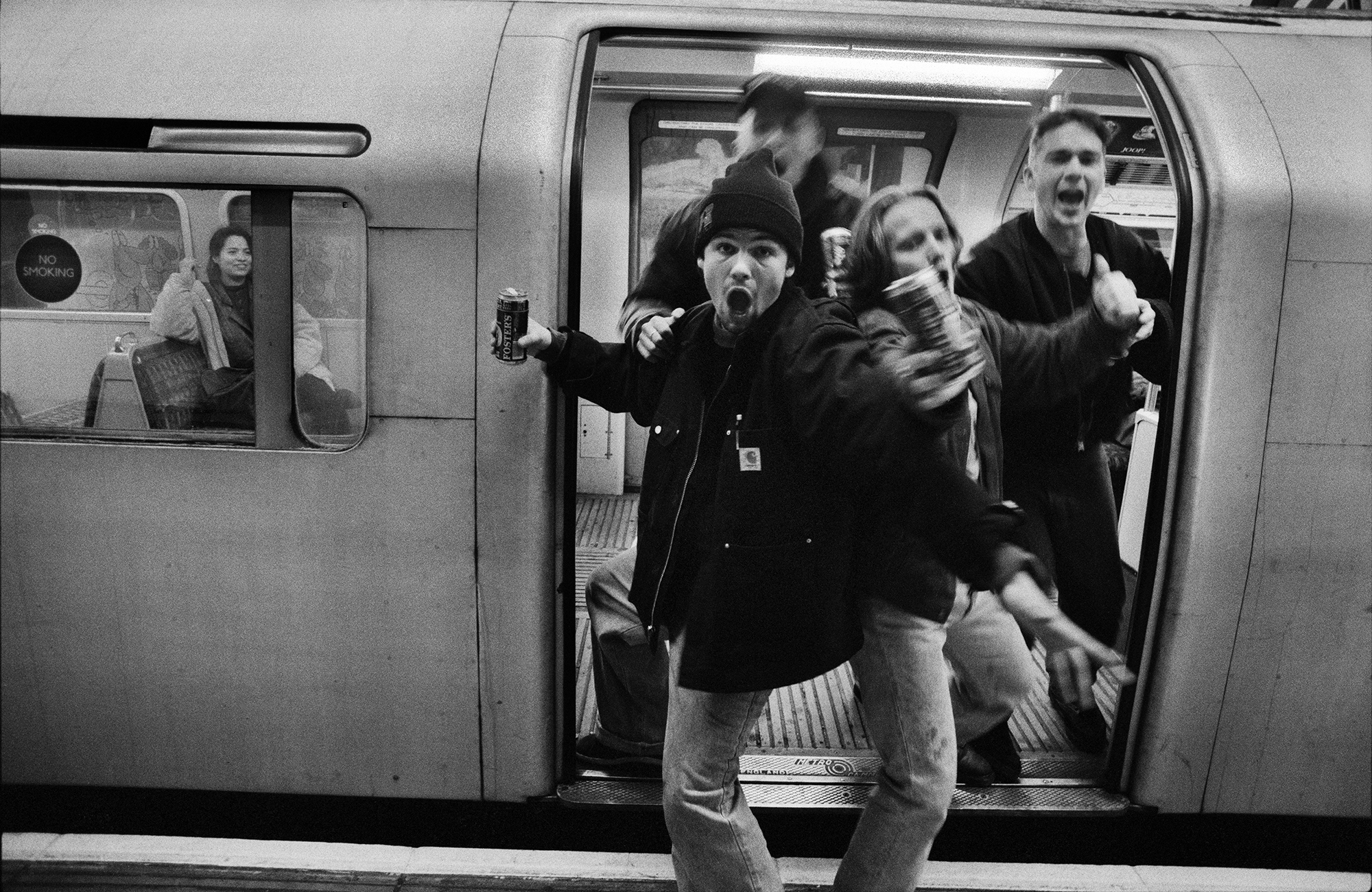 Black and white image of crowded train carriage with passengers pressed together in doorway, some appearing to push or fall out.