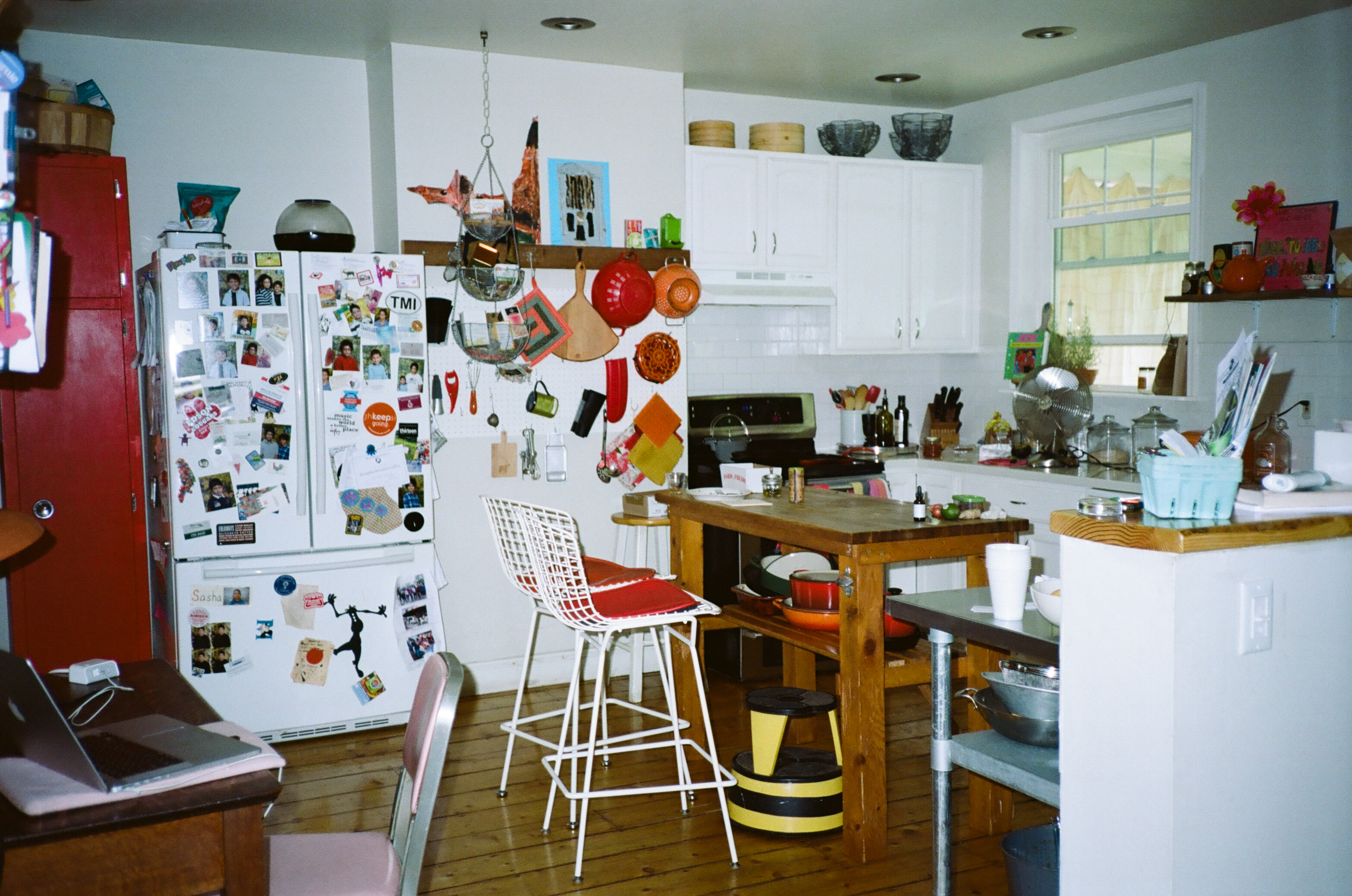 Kitchen with white units, wooden worktops, red fridge covered in photos/magnets, white wire bar stool, and shelving with various items.