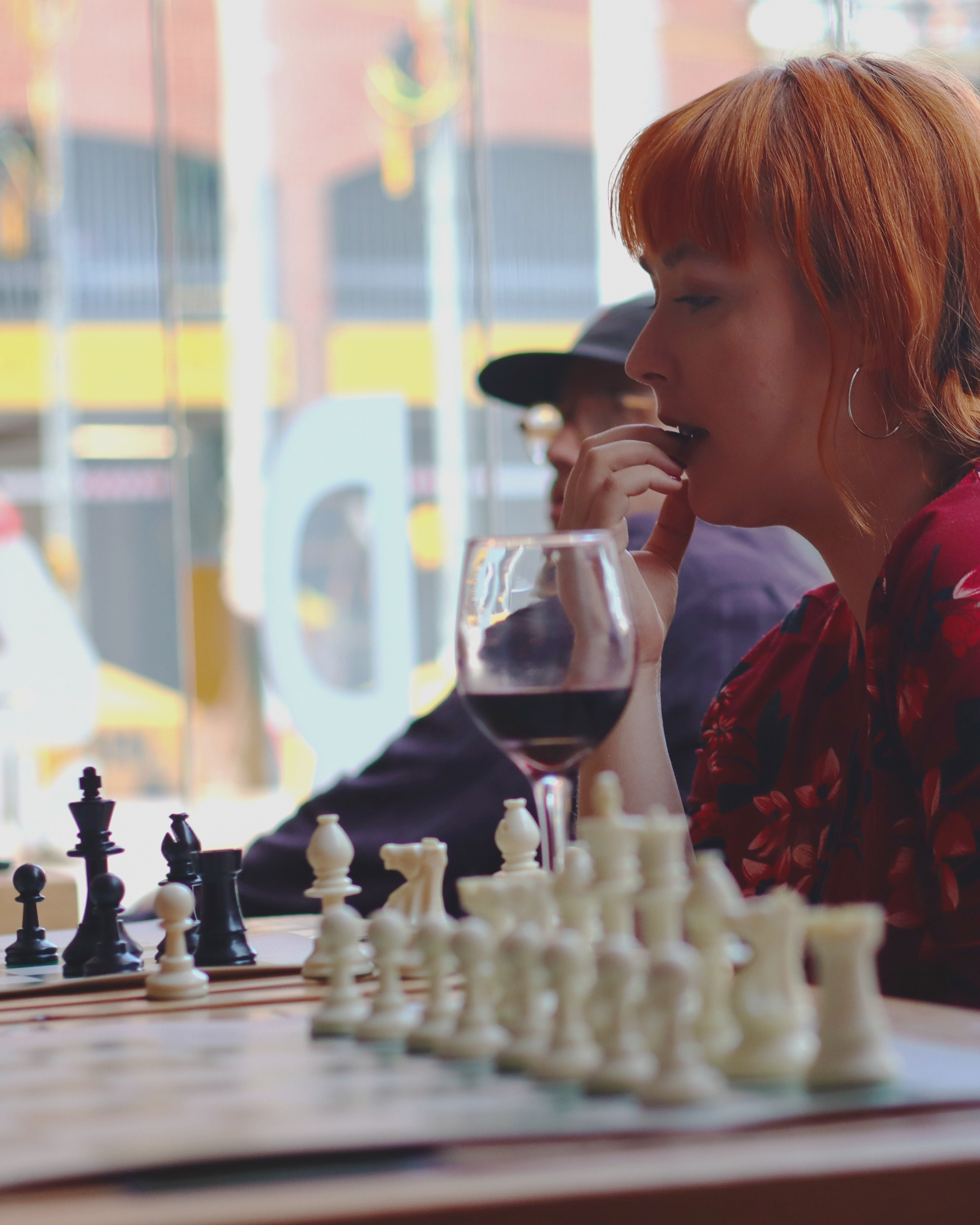 Woman with red hair and fringe sits at chess board, holding red wine glass. Blurred colourful buildings visible through window behind.