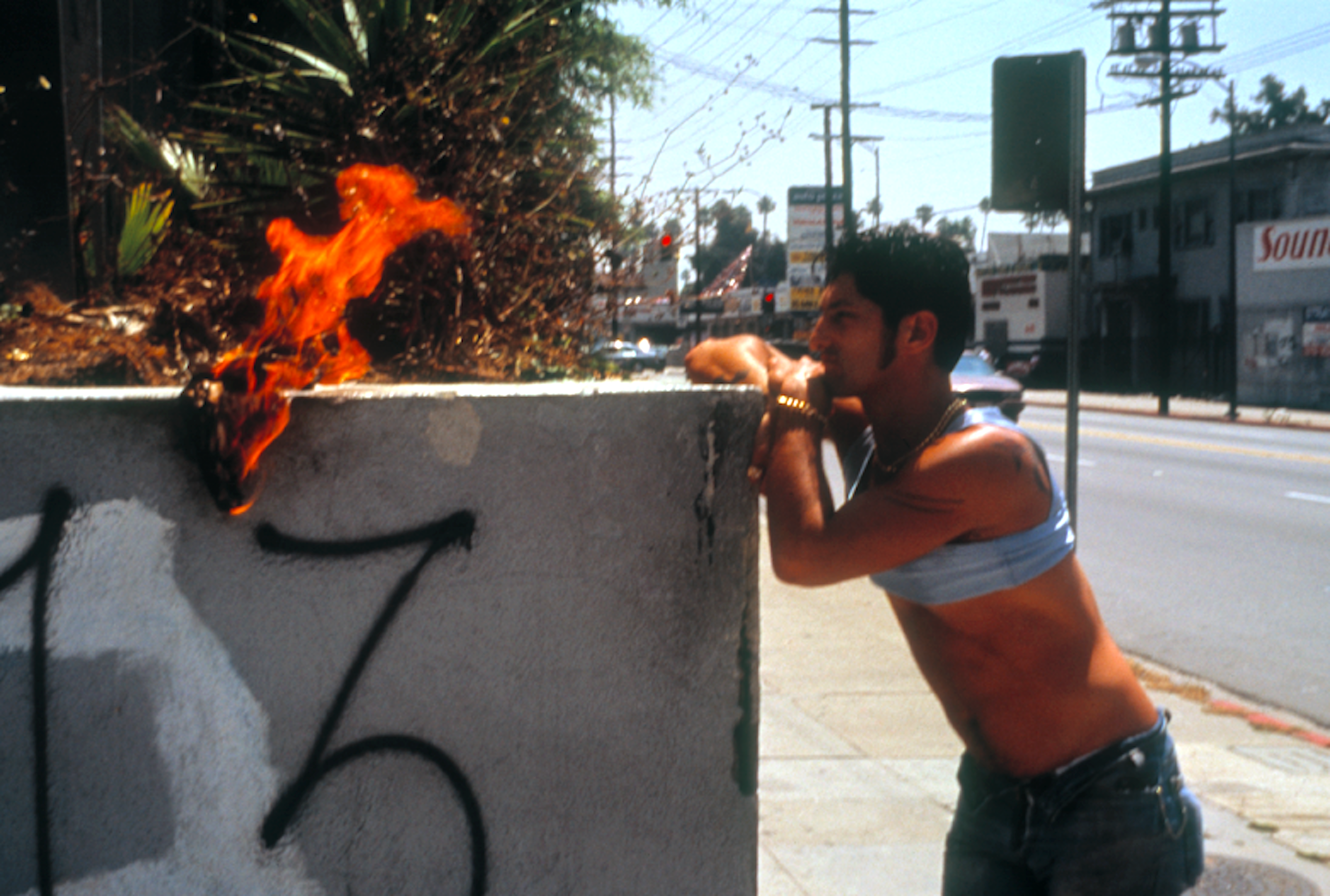 Man kneeling beside concrete barrier with orange flames emerging from top, street scene with power lines and buildings in background.