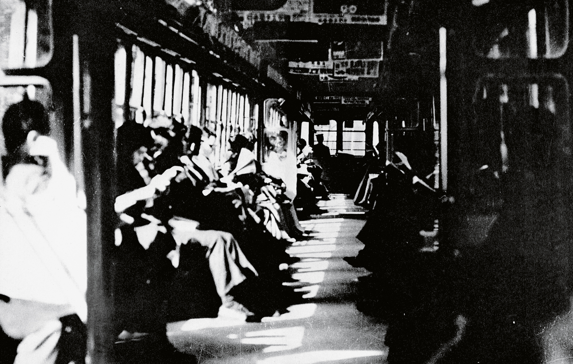 Black and white image showing interior of vintage train carriage with passengers seated along both sides, windows visible on left wall.