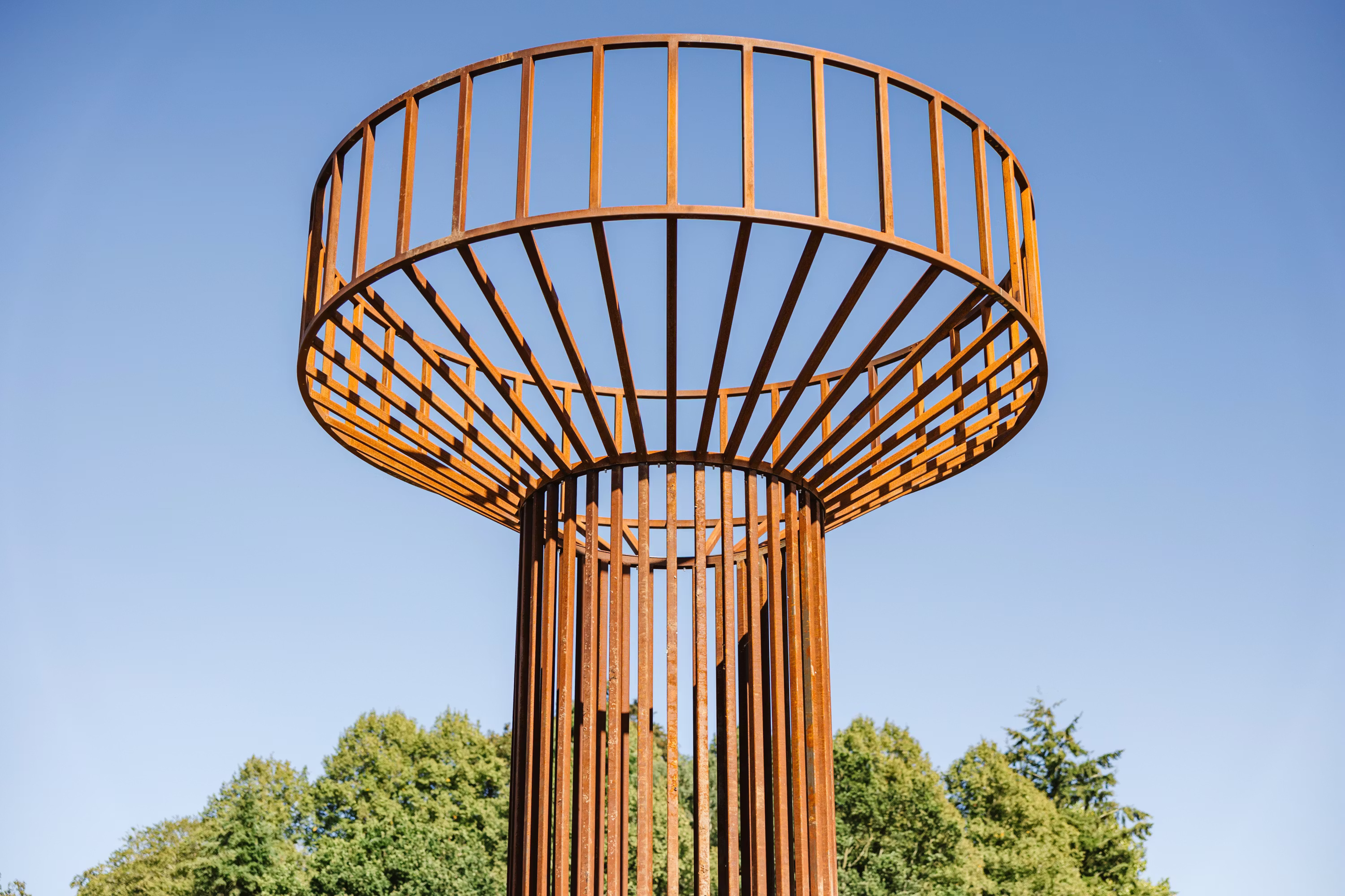 Modern sculptural tower with cylindrical orange metal framework topped by circular canopy structure against blue sky and green trees.