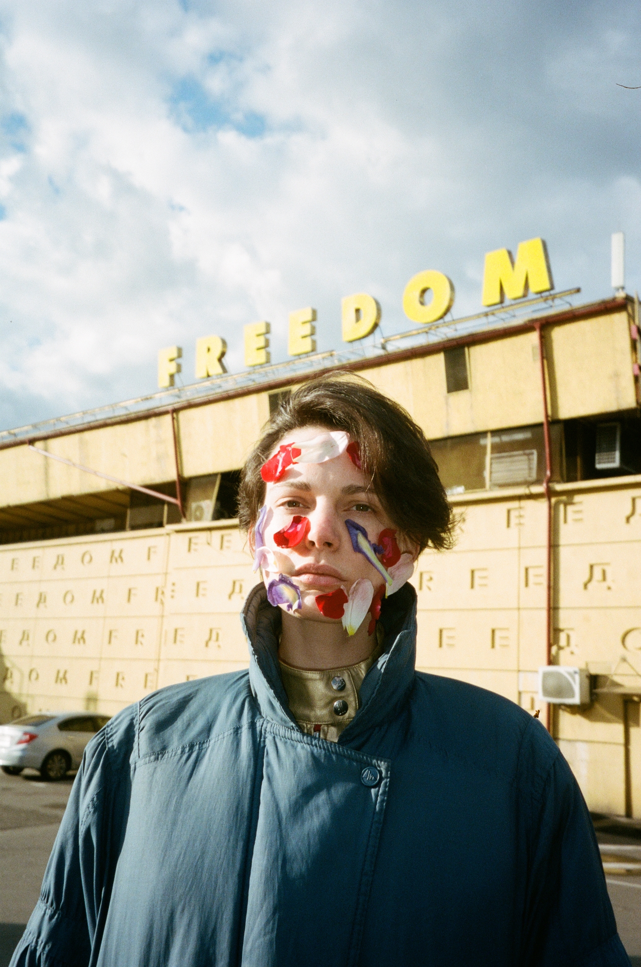 A person with painted face and an expression of concern or distress, standing in front of a building with the word "Freedom" displayed on its facade.