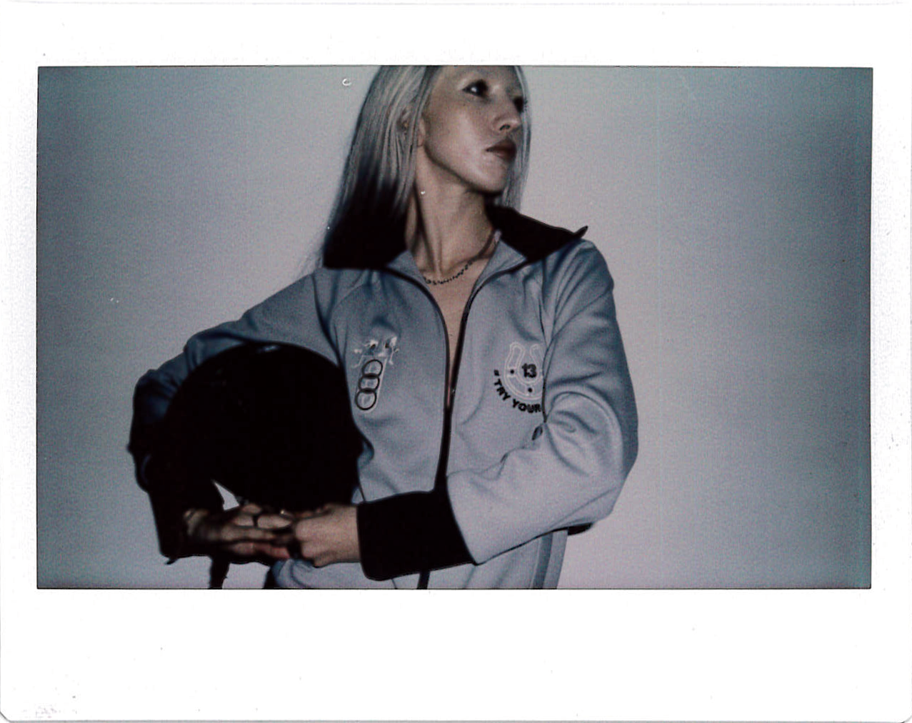 A woman with long blonde hair wearing a grey jacket and black accessories.