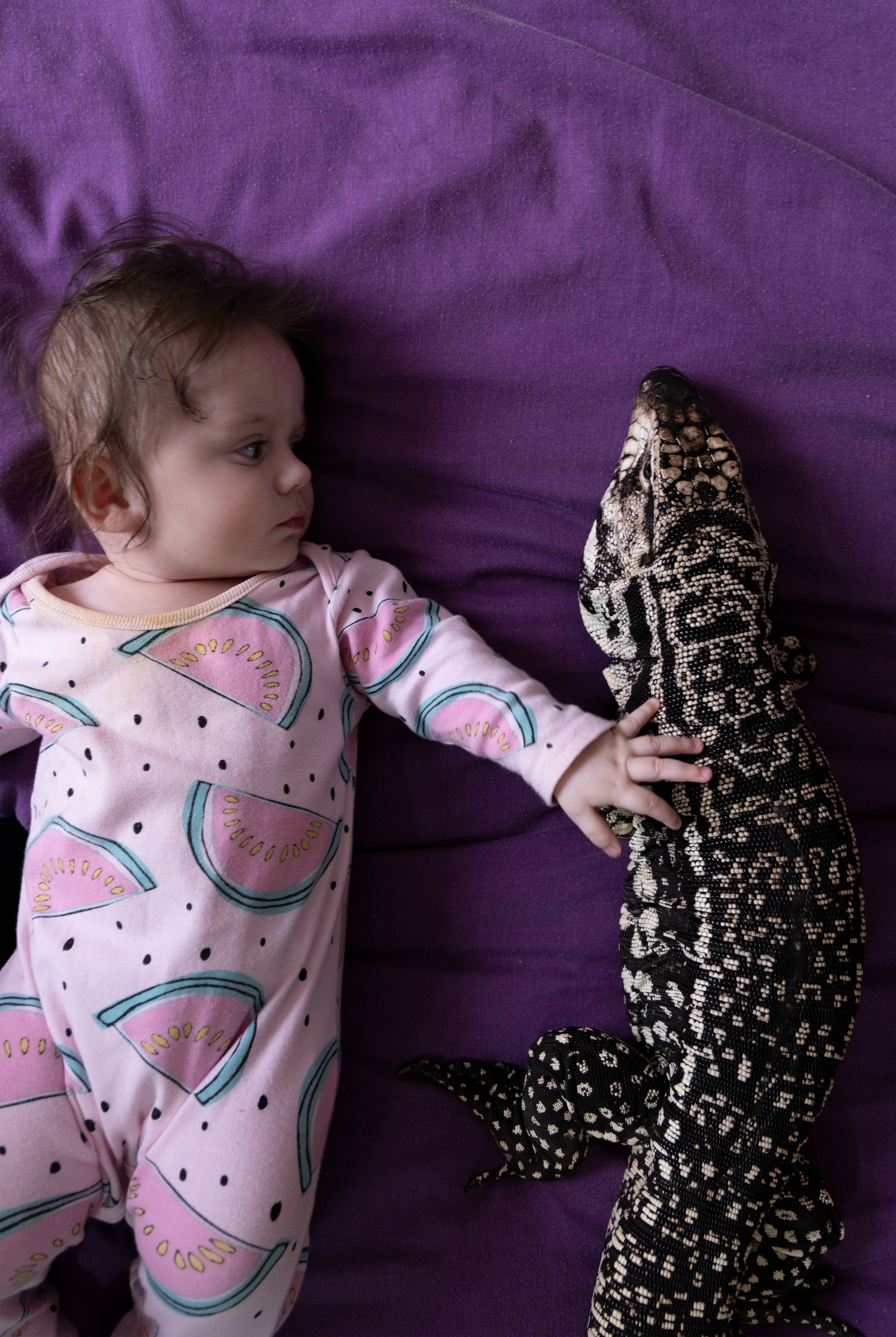 Infant in patterned pink sleepsuit gazing at patterned plush toy on purple sheet.