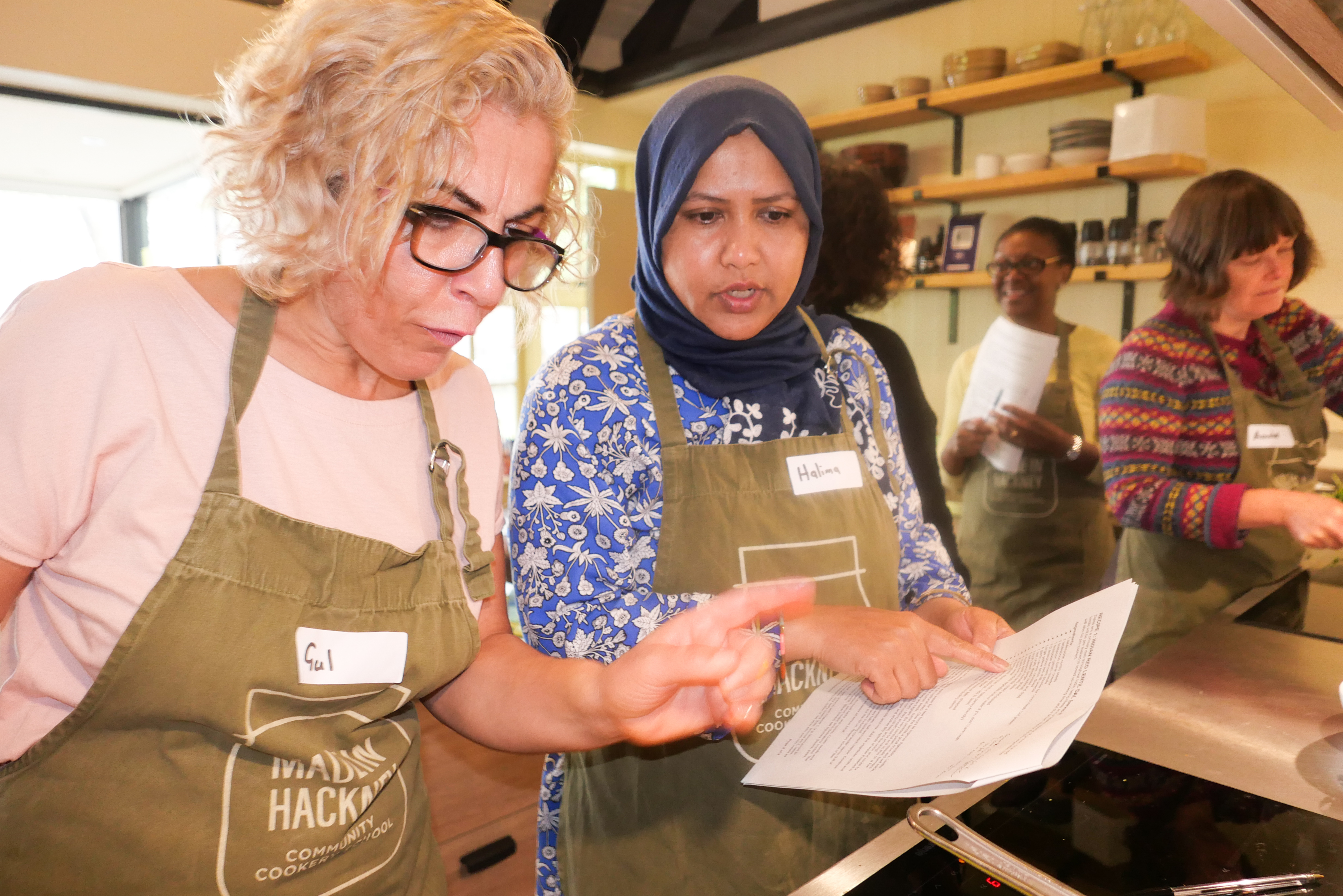 Two women in green aprons examining papers in kitchen. Blonde woman with glasses on left, woman in blue headscarf on right.