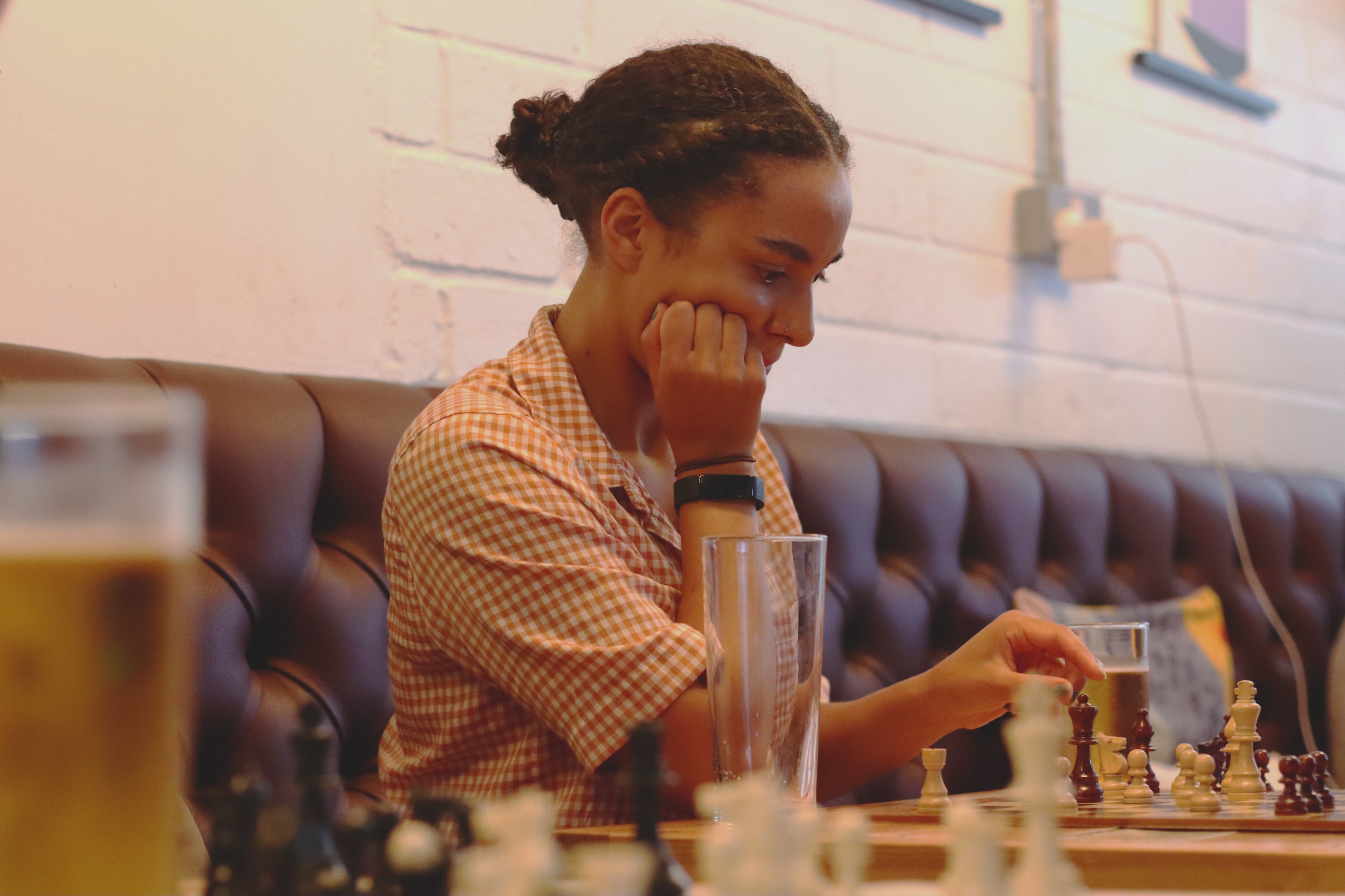 Woman in checked shirt playing chess at table with glass, seated on blue banquette seating in restaurant or café setting.