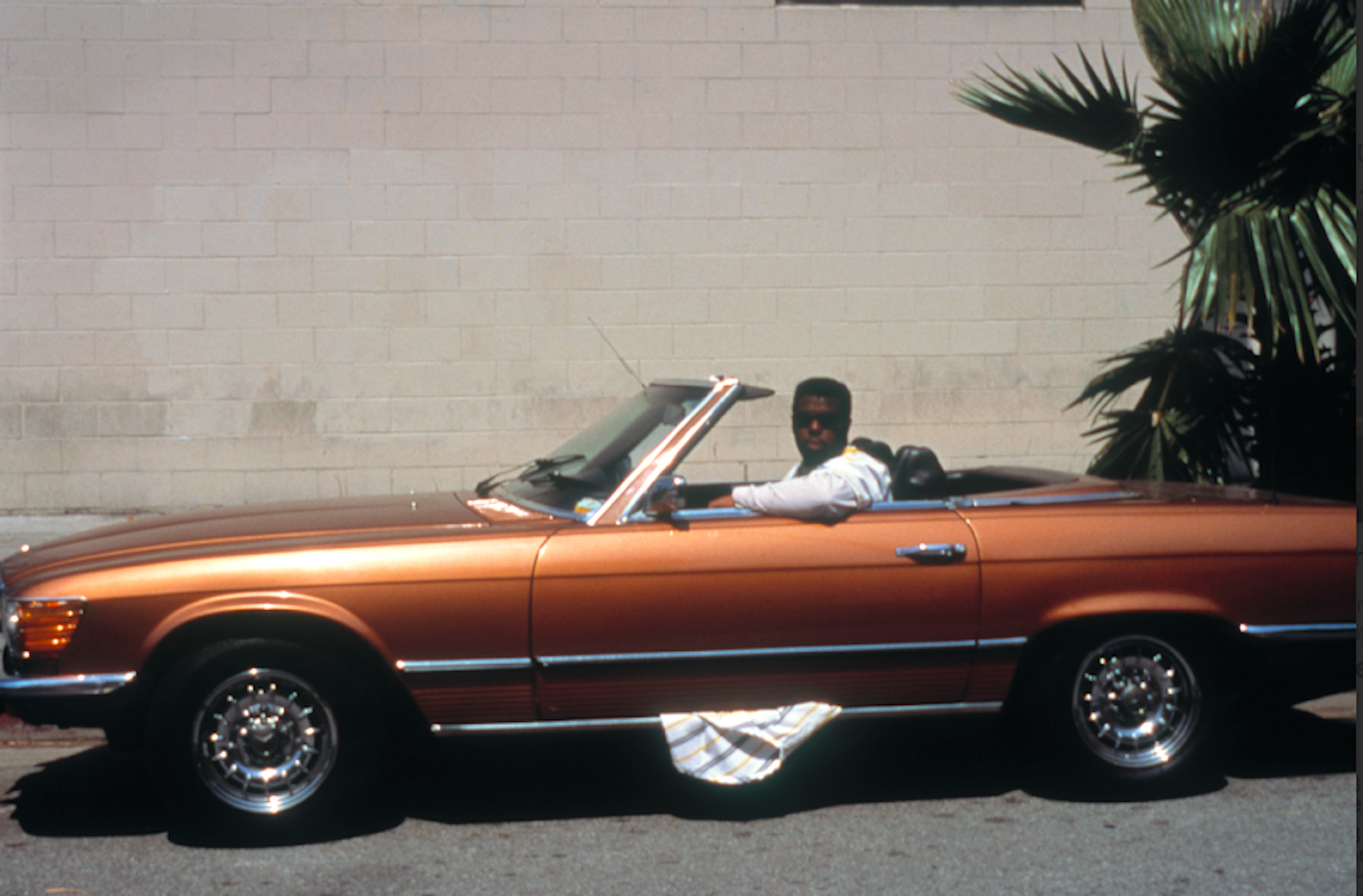 Orange convertible car parked beside white wall with palm tree. Driver in white shirt seated inside vehicle.