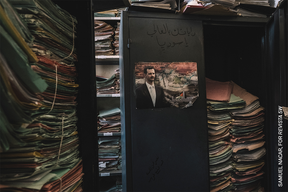 Stacks of files and documents, a portrait photograph of a man in a suit visible on a blackboard.