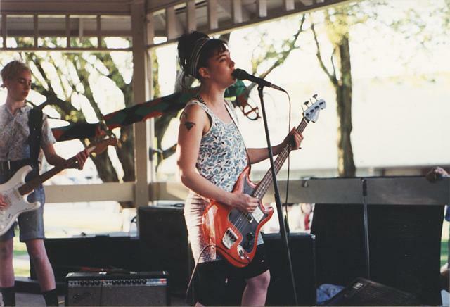 Person in patterned top playing electric guitar on stage with band members in background.