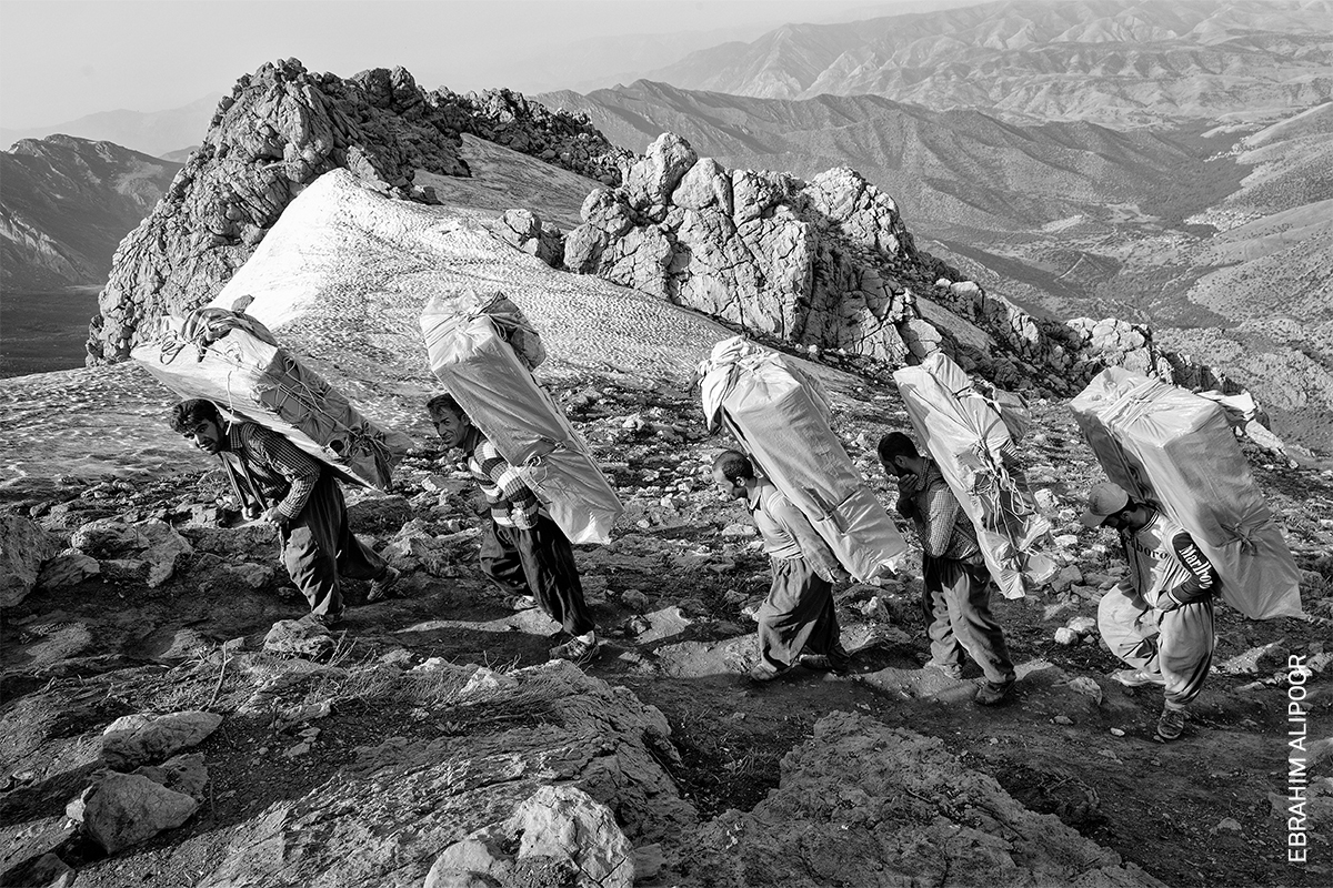 Black and white image of people carrying materials up rocky, mountainous terrain.