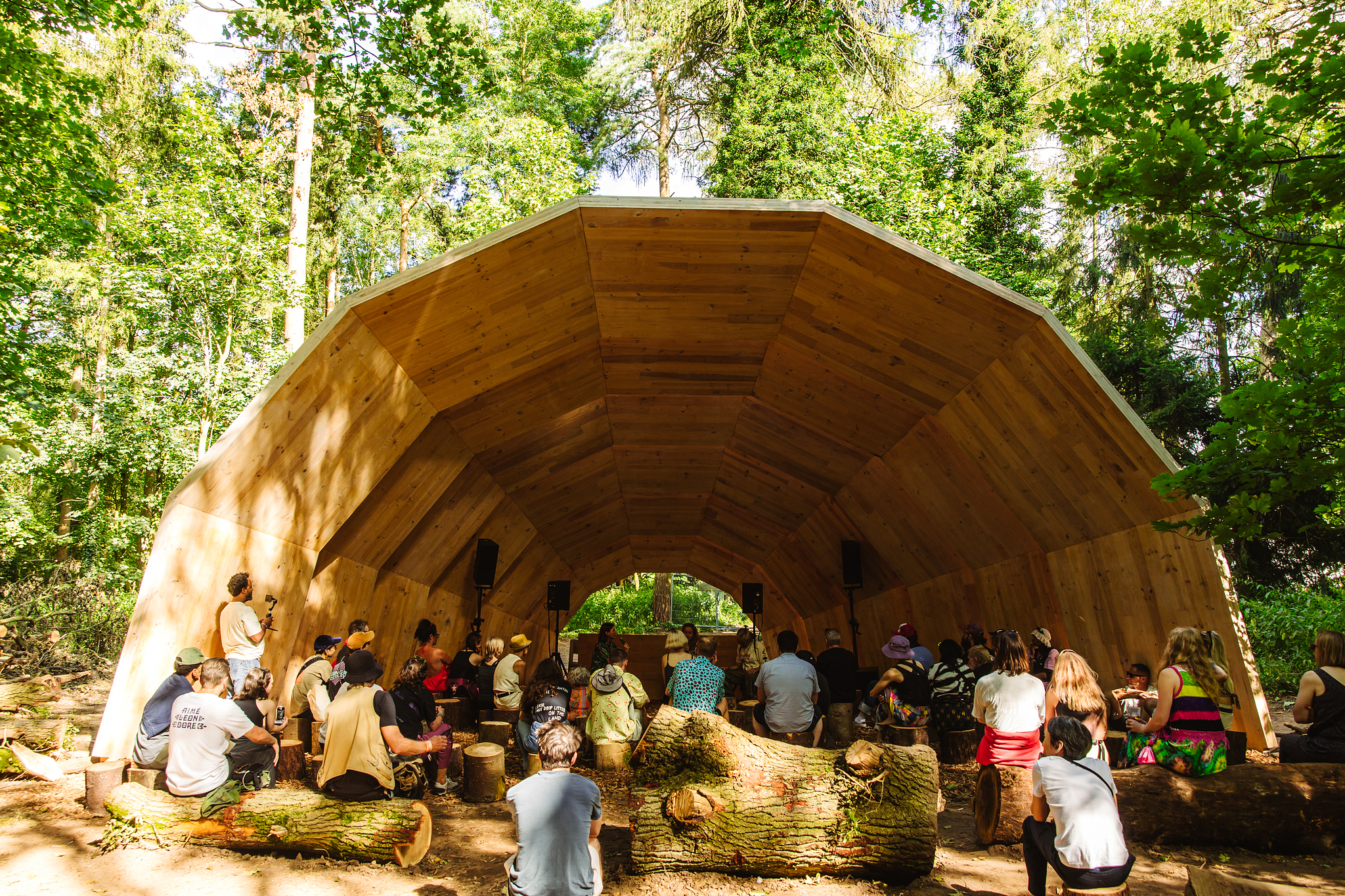 Large wooden amphitheatre with curved roof in forest setting. Audience seated on log benches facing stage area surrounded by green trees.