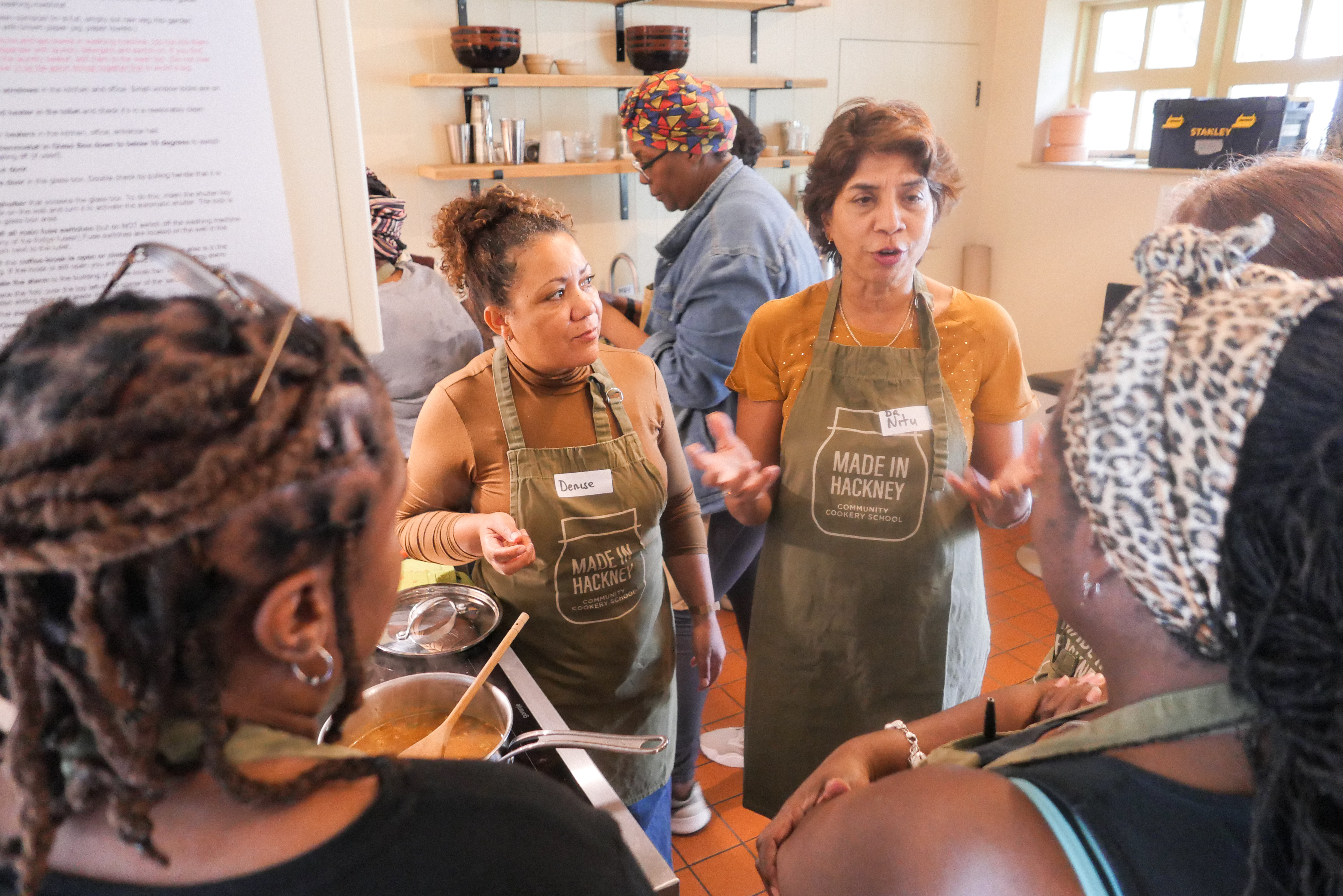 Two women in green aprons leading cooking class in bright kitchen with participants seated at counter, shelves and windows visible