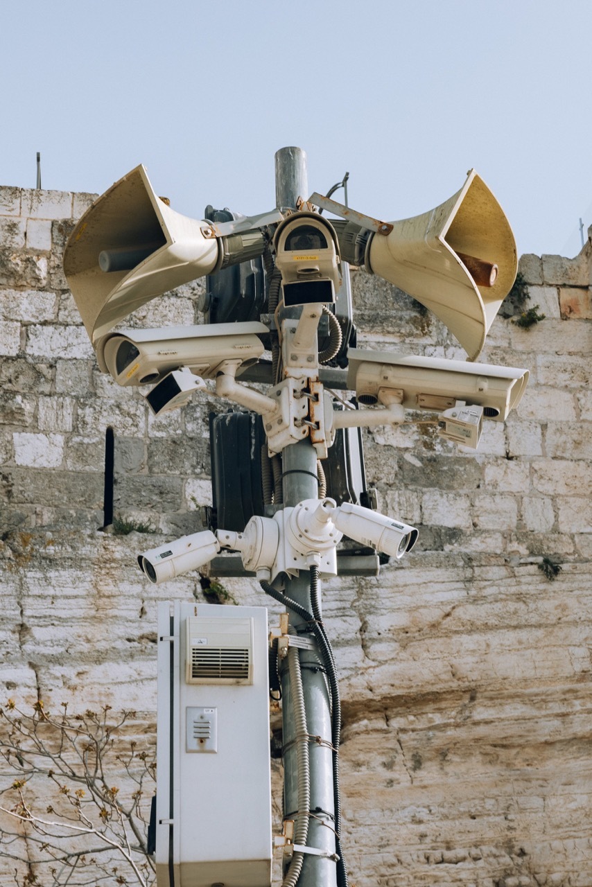 Unusual outdoor audio equipment with multiple horn speakers and a cylindrical body against a weathered stone wall.