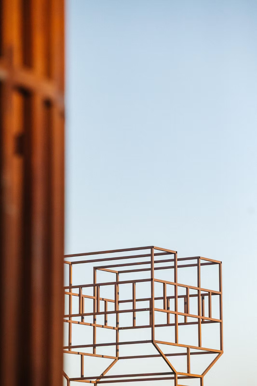 Orange metal geometric cube framework structure against pale blue sky, with blurred orange column on left side.