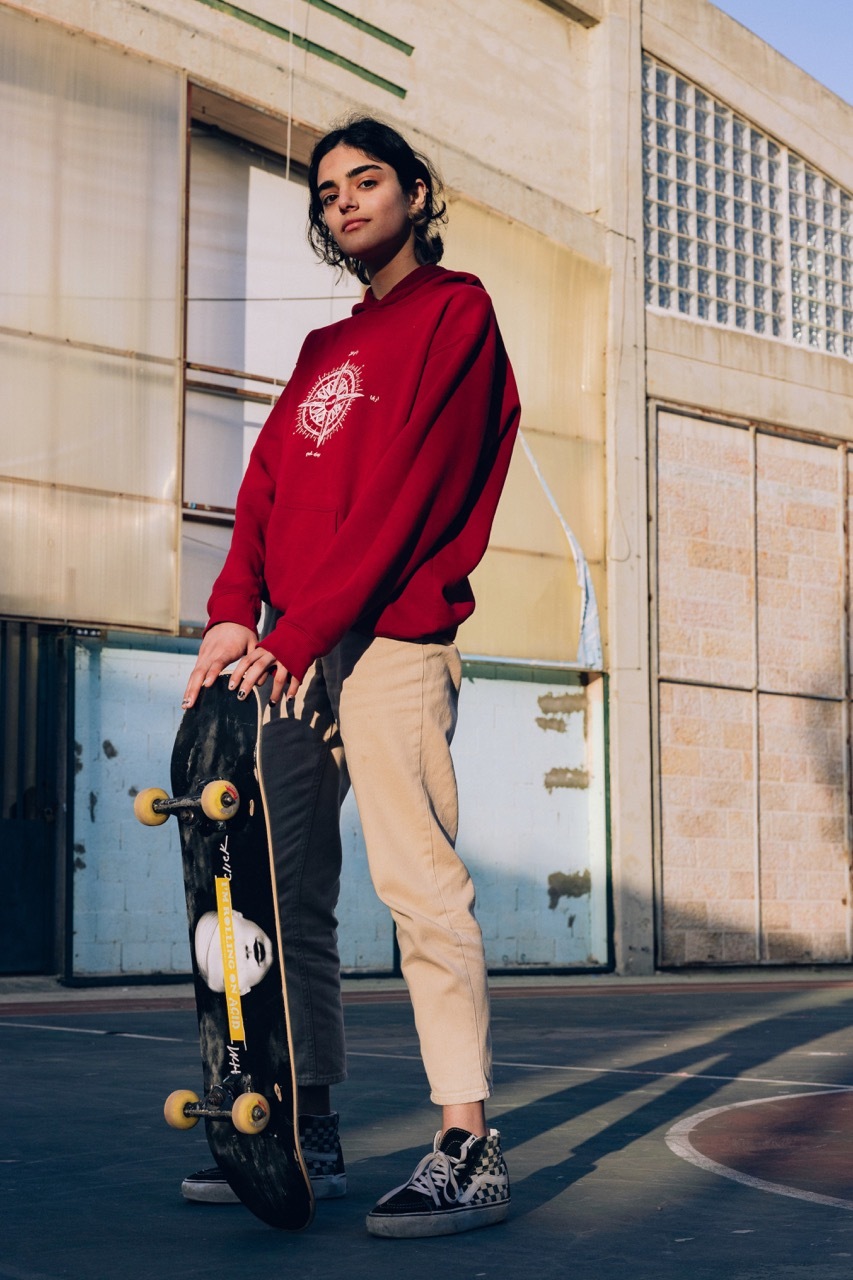 Young woman in red sweatshirt and casual trousers standing with skateboard.