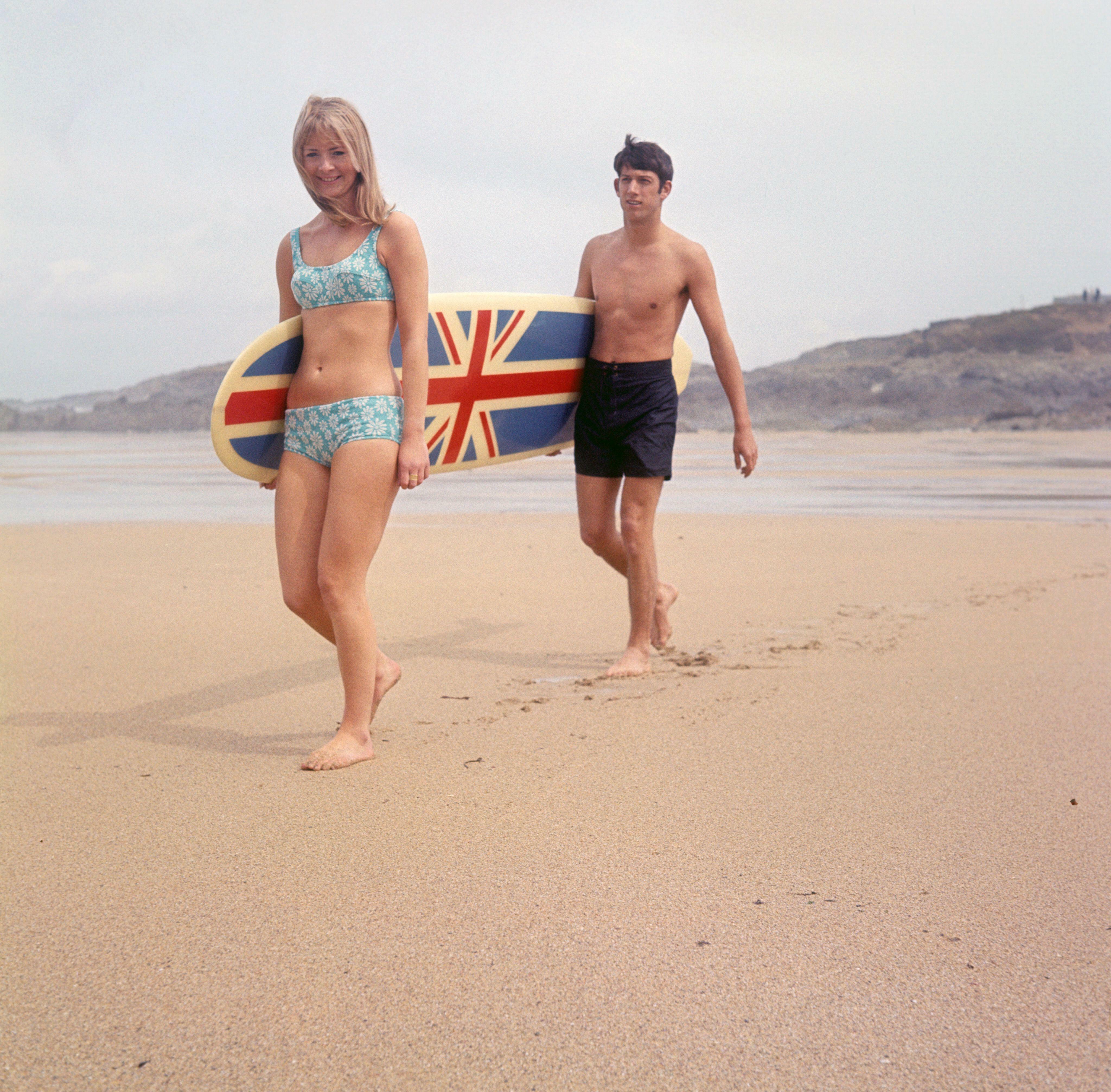 Man and woman in swimwear walking on beach, carrying Union Jack flag.
