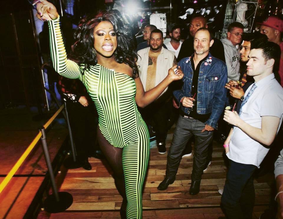 A woman in a green and white striped jumpsuit dancing on a stage with a crowd of people behind her.