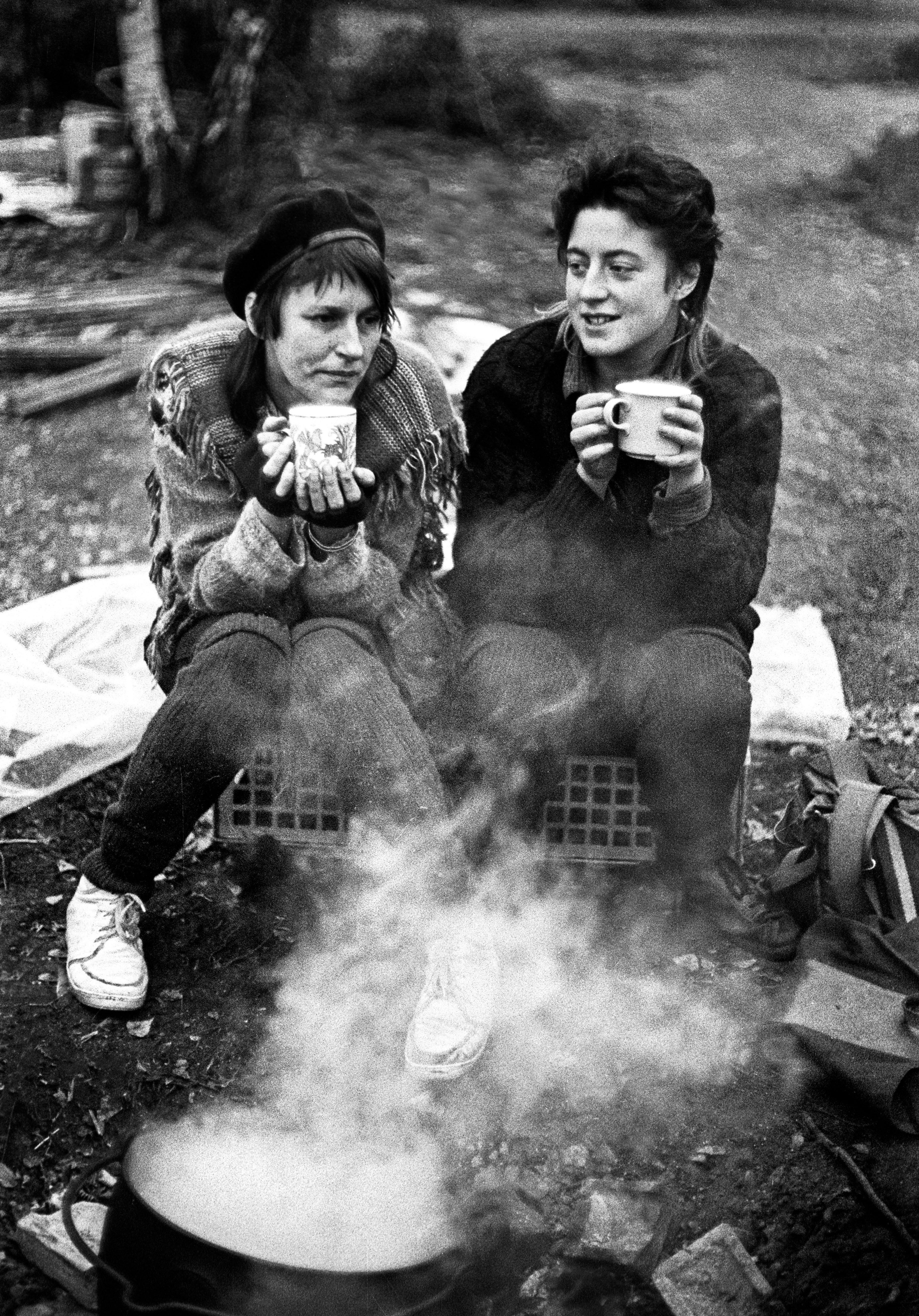 Two women sitting outdoors holding mugs, with steam rising from large pot in foreground. Black and white photograph.