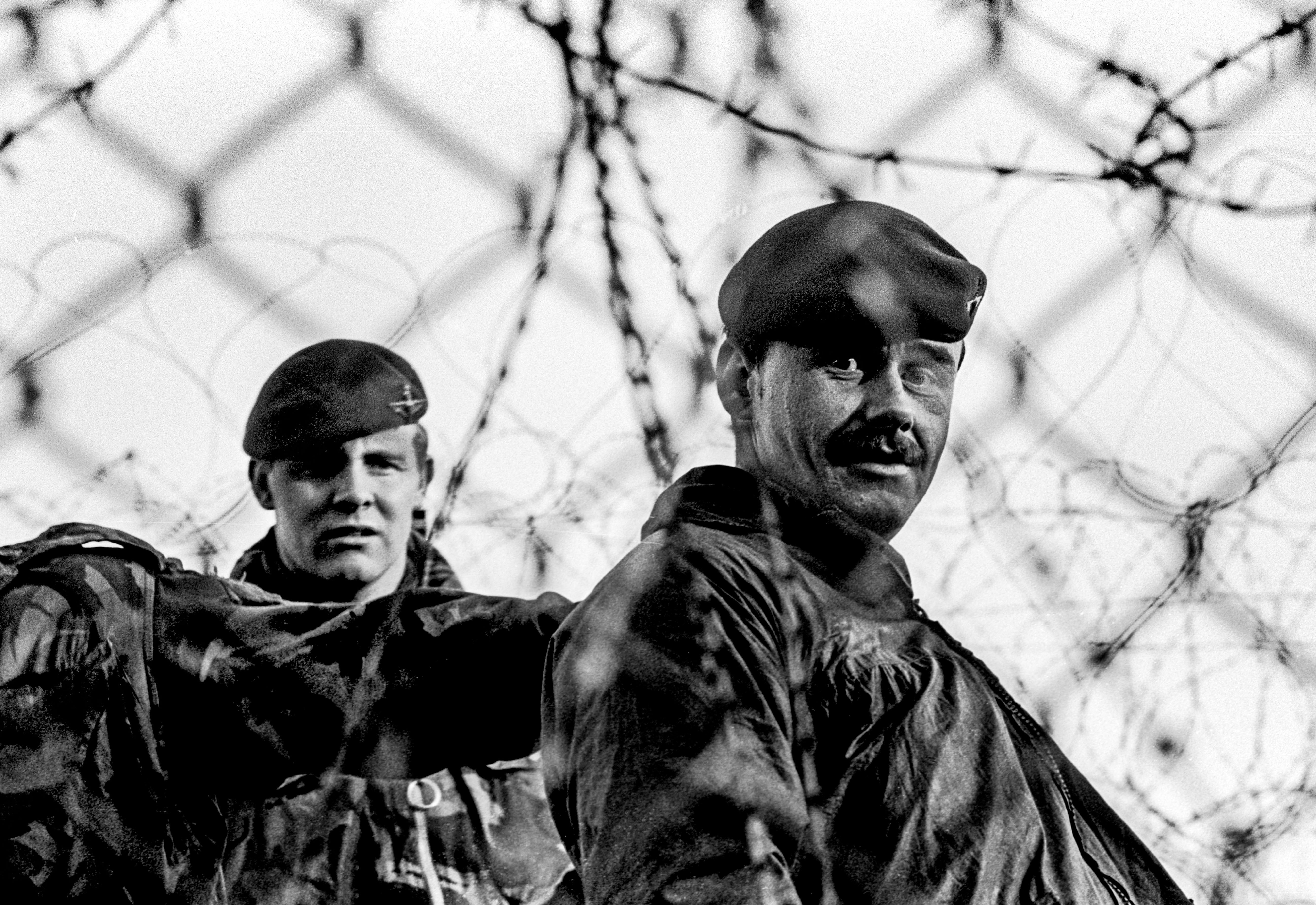 Two soldiers in military uniforms and berets standing behind barbed wire fence, black and white photograph.