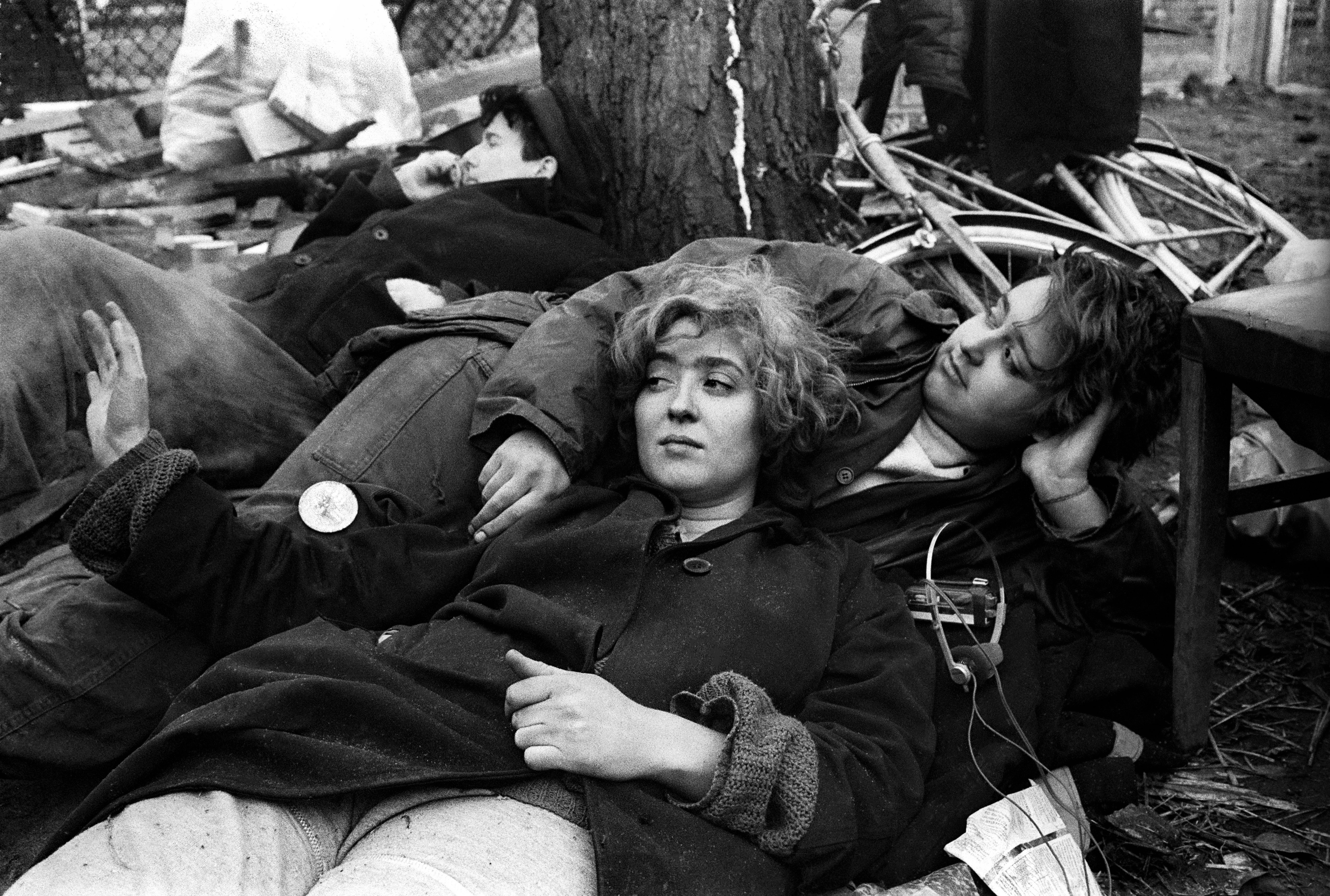 Black and white image of two people lying on ground amongst debris and scattered papers, with tree trunk visible in background.