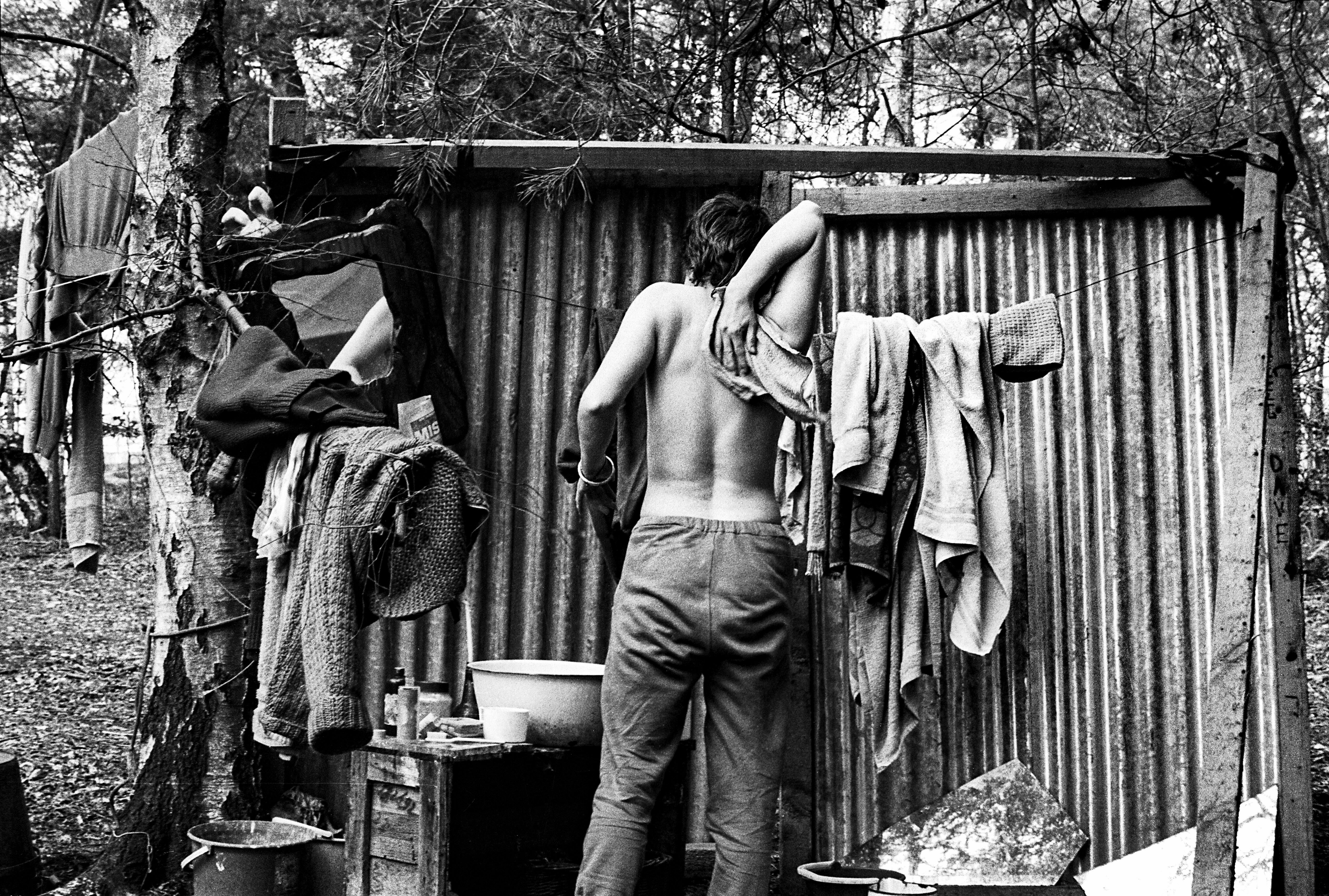 Shirtless man washing at corrugated metal shelter with clothes hanging on line, surrounded by trees in black and white image.