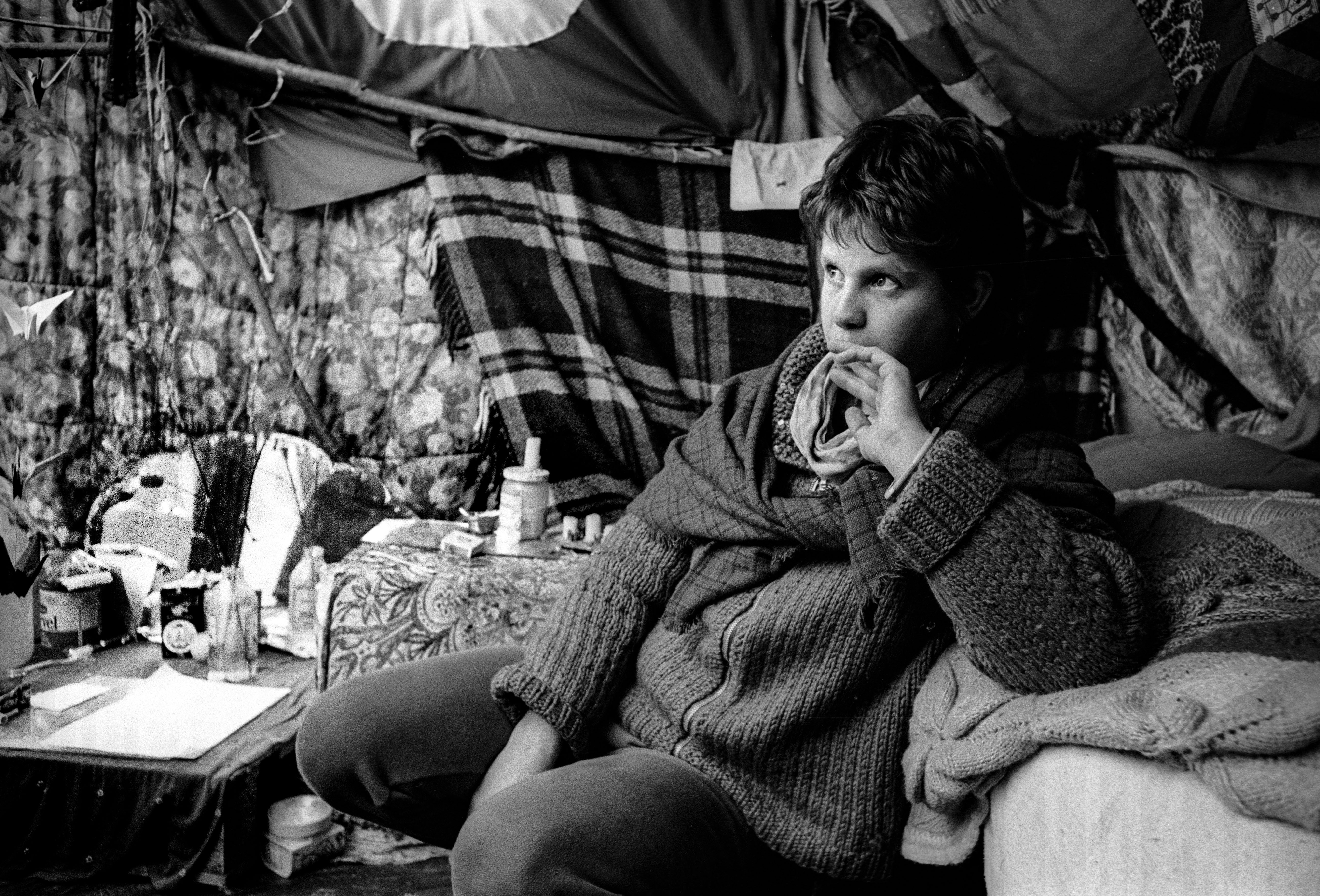 Black and white image of person sitting on bed in cramped room, surrounded by hanging fabrics, small table with bottles visible.