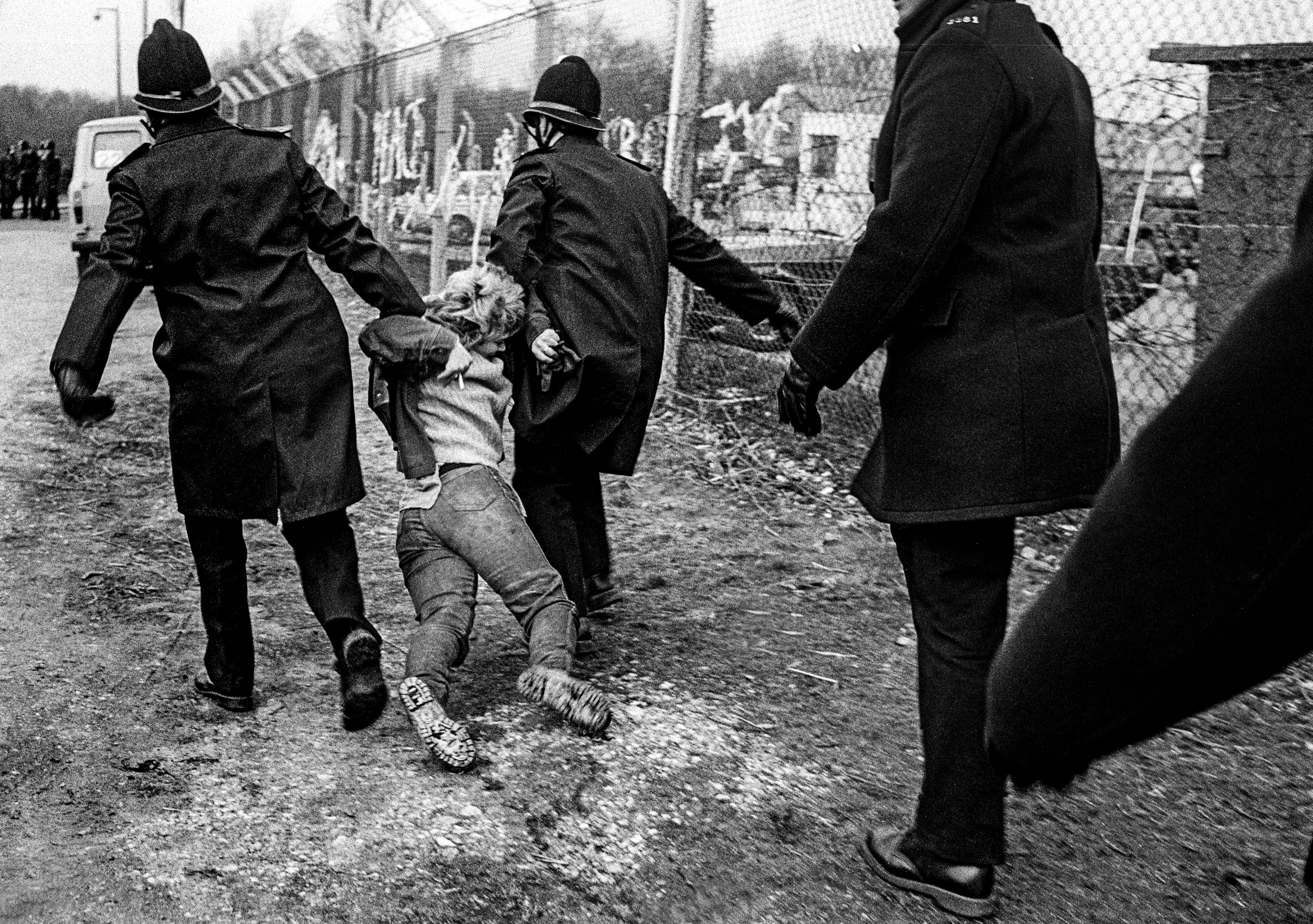 Police officers dragging a person across rough ground near chain-link fencing and damaged buildings in black and white.