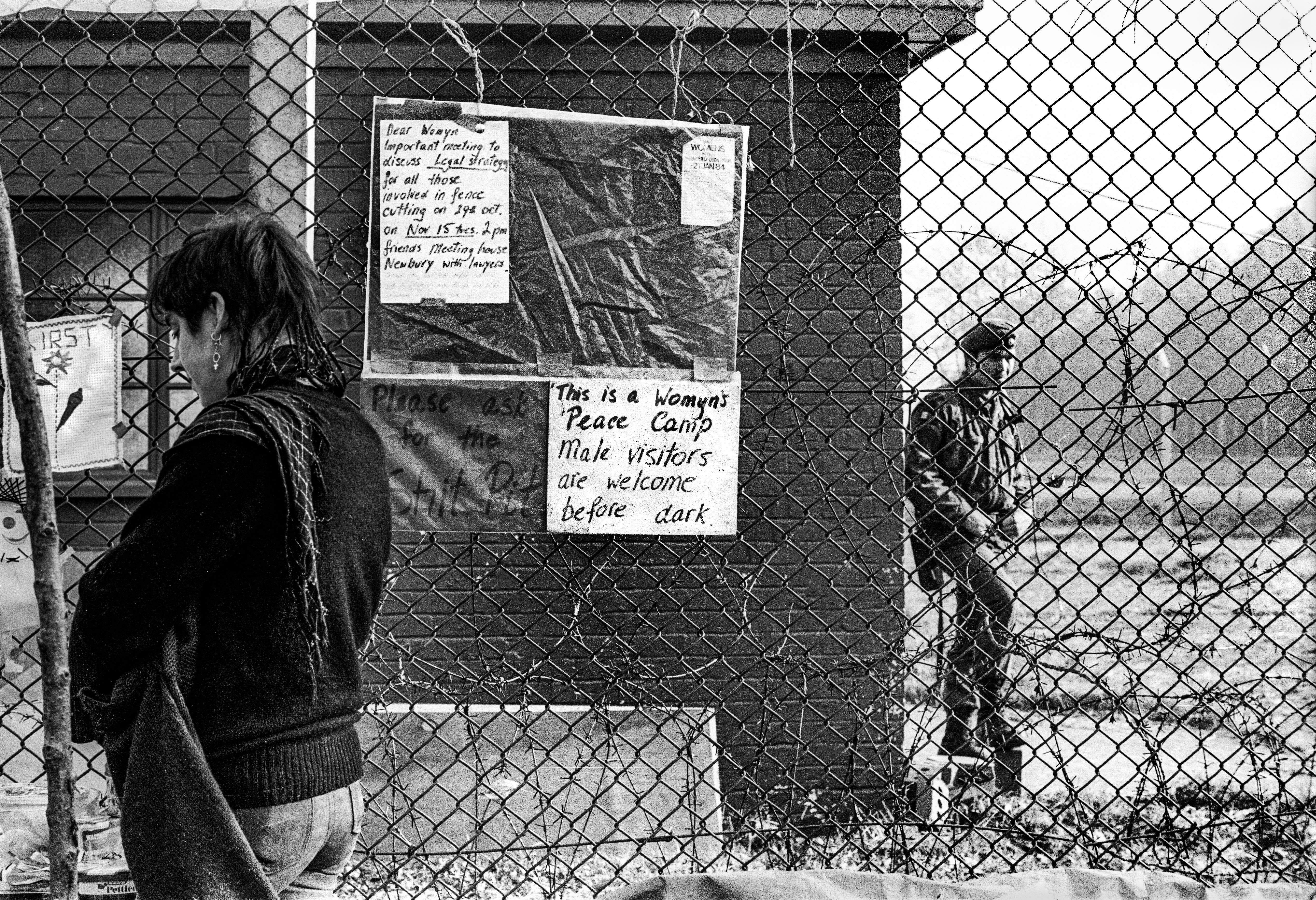 Black and white image showing chain-link fence with posted notices, woman walking left, uniformed figure visible through fence on right.