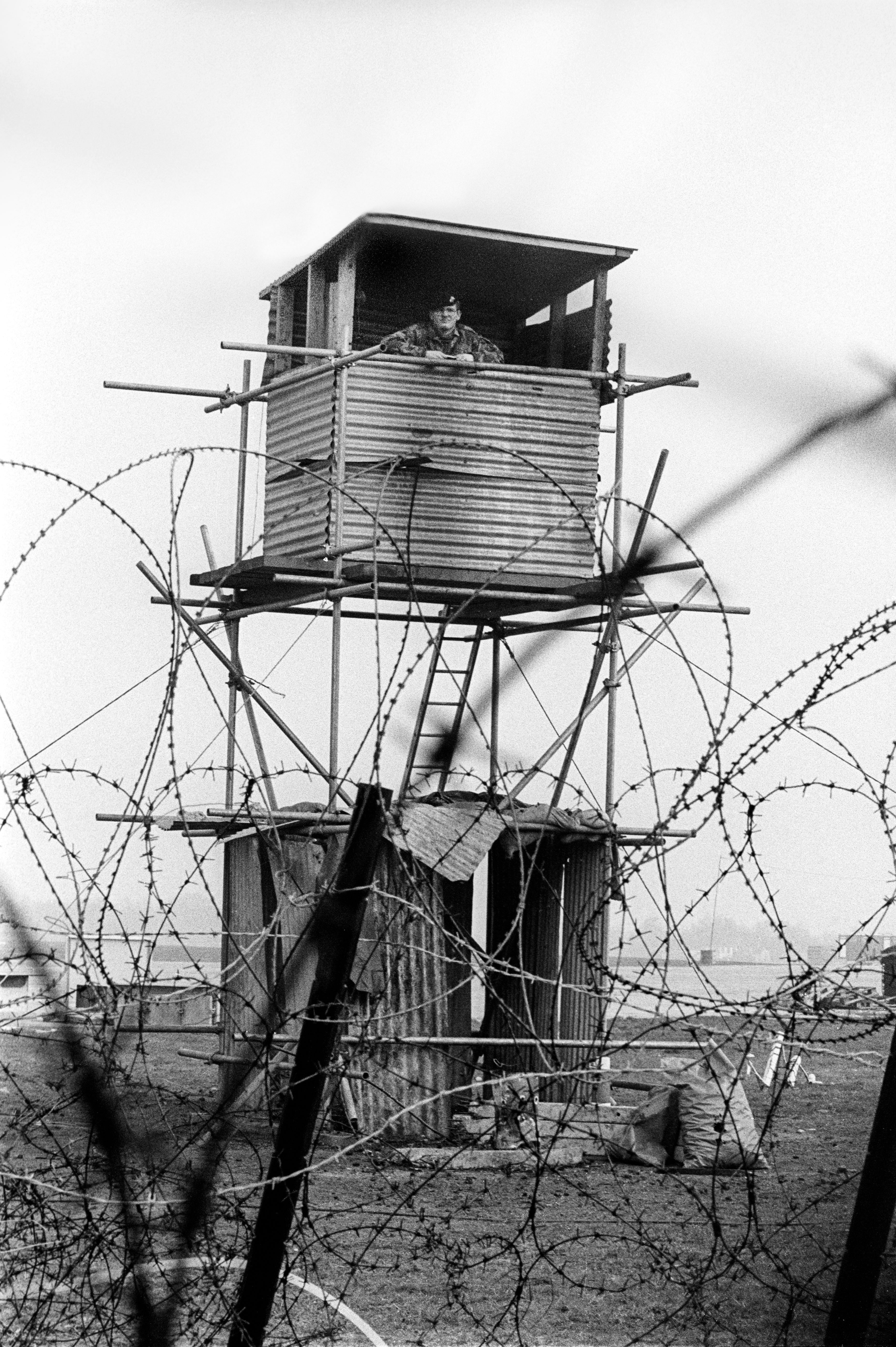 Military watchtower with guard, surrounded by coiled barbed wire fencing. Black and white photograph.