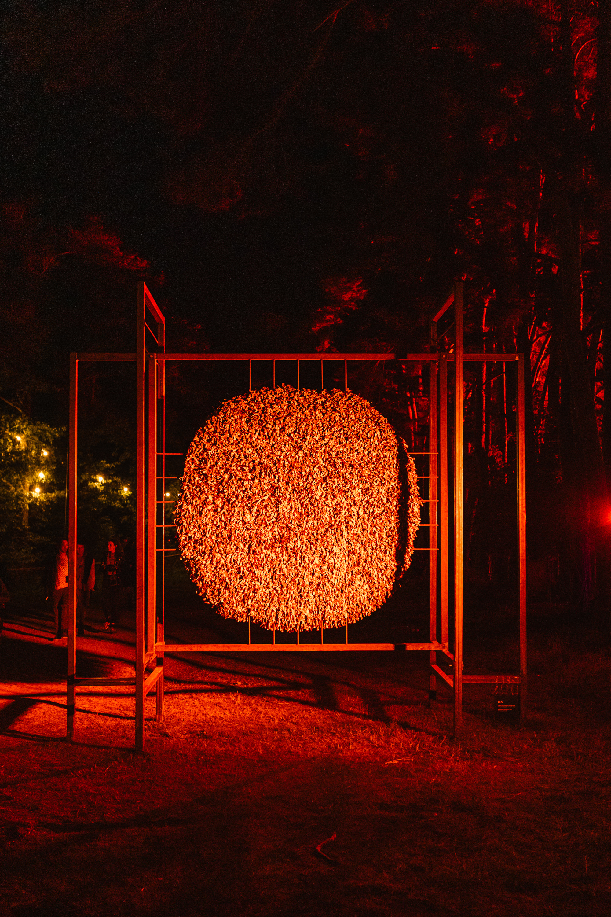 Illuminated orange circular sculpture suspended within red metal frame structure at night, surrounded by trees and warm lighting.
