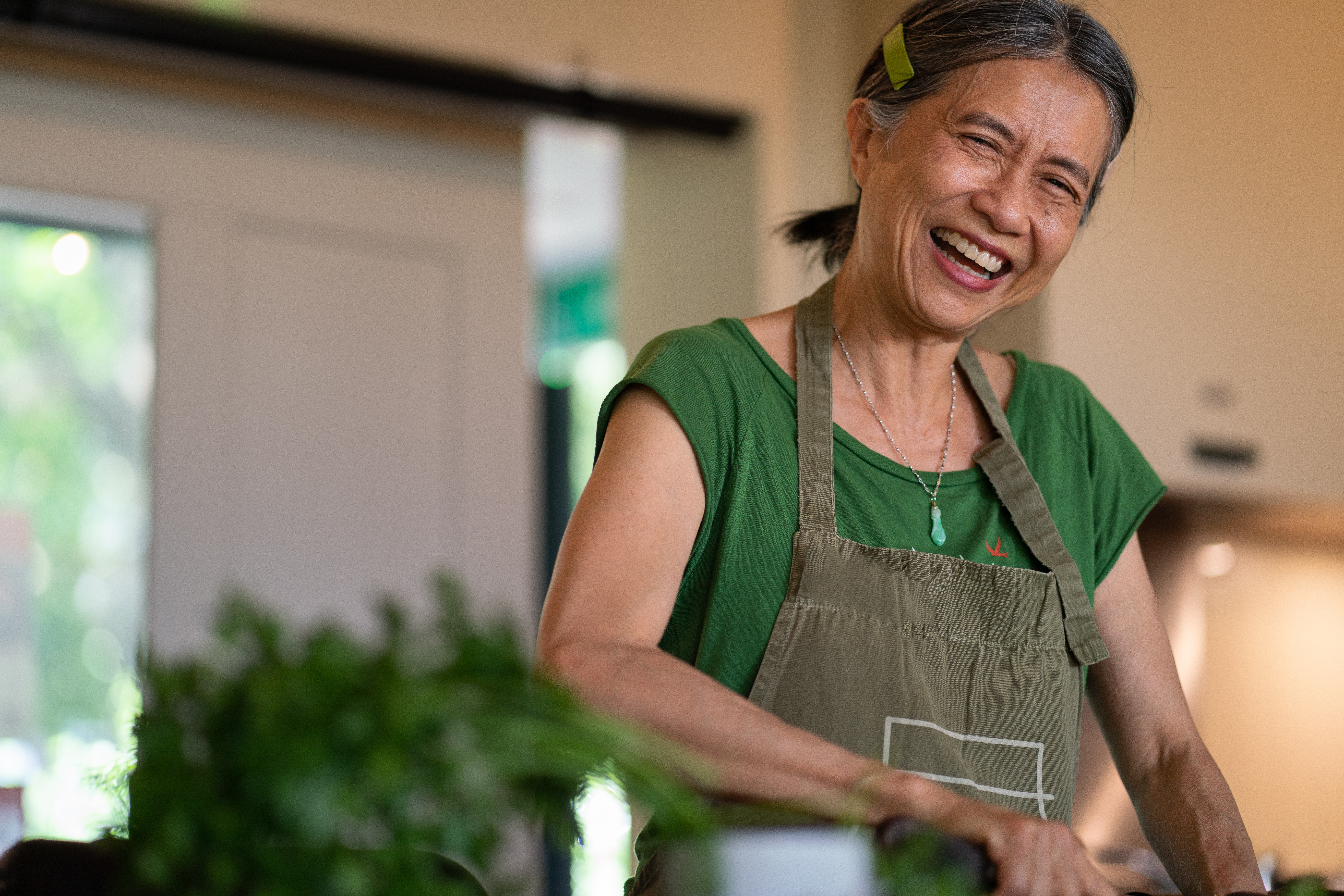 Woman with grey hair in green shirt and beige apron laughing whilst working in kitchen with plants visible in background.