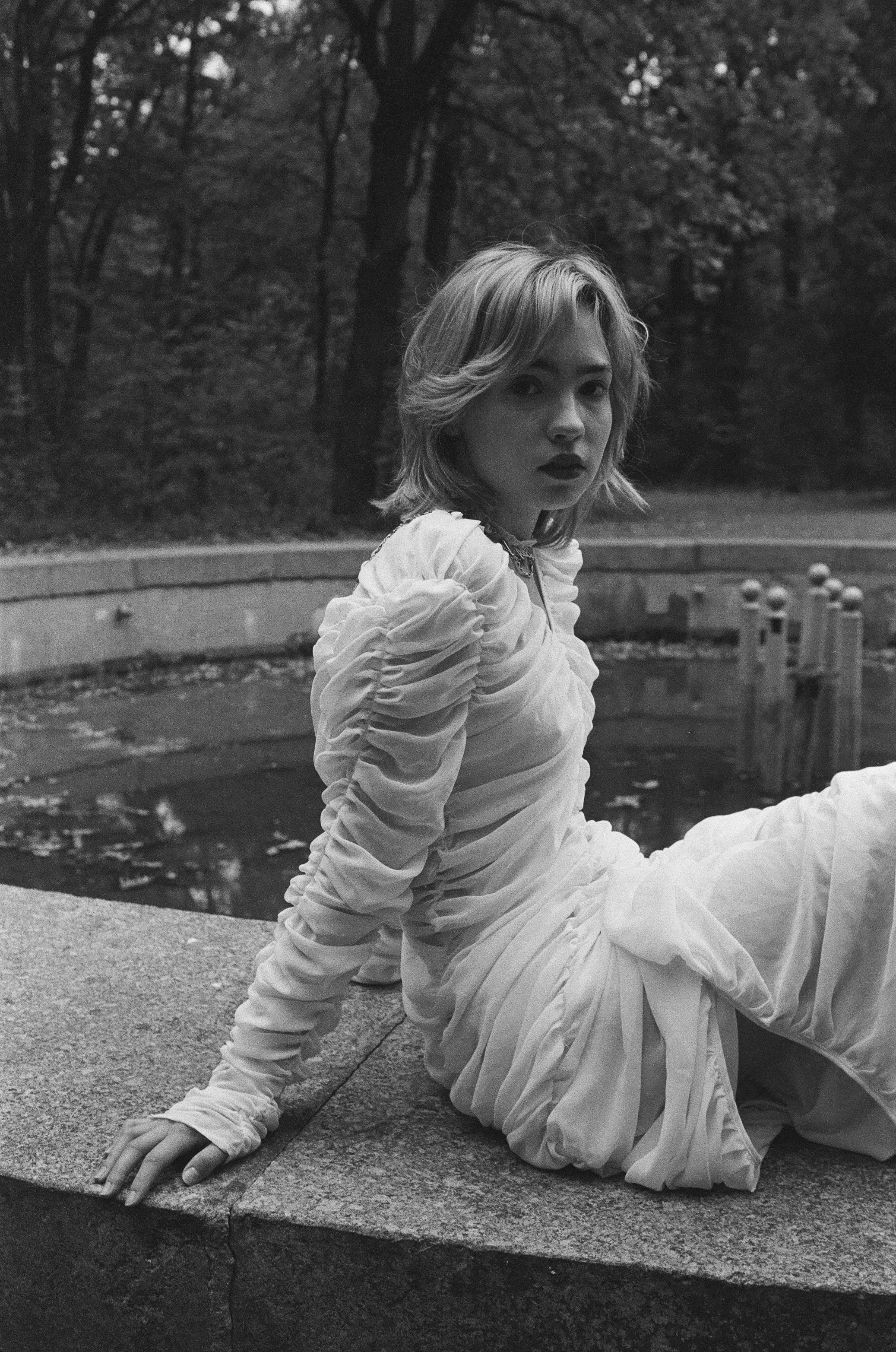 Woman in a white ruched dress sitting by a fountain in a wooded area, black and white photograph.