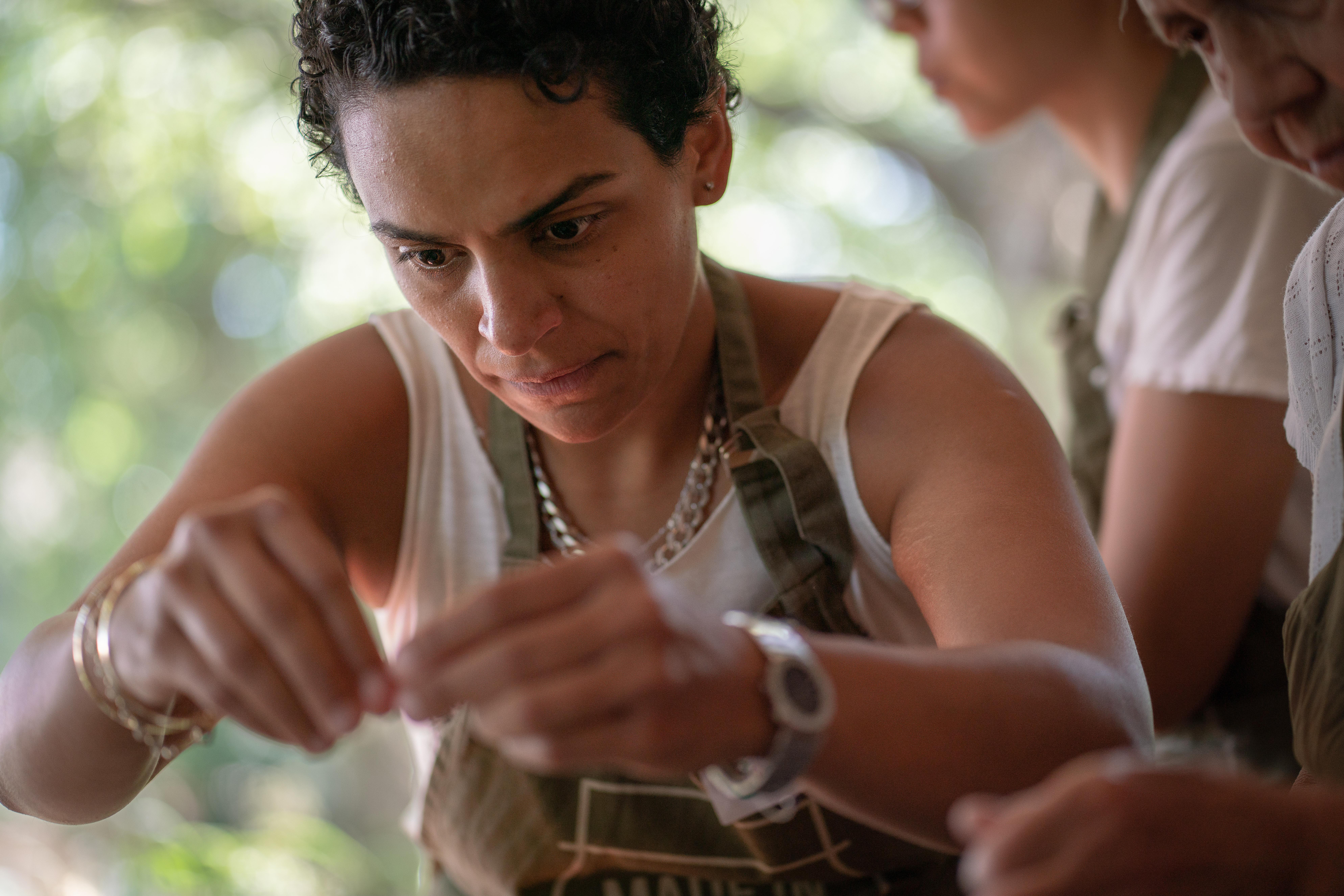 Woman with curly dark hair in white vest top handling white object, another person partially visible on right, blurred green background.