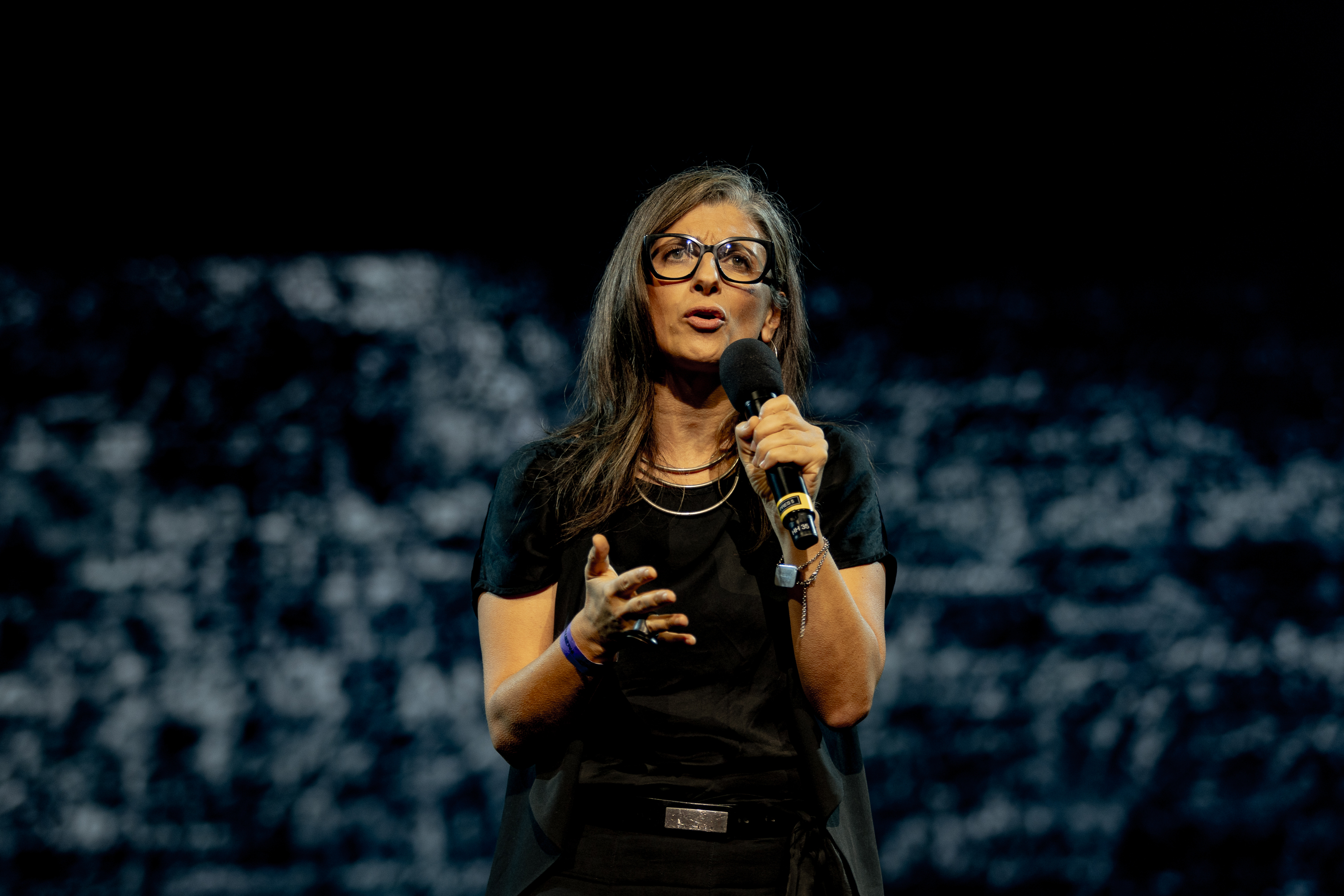 Woman with glasses and long brown hair speaks into microphone, wearing black top against blurred dark background.