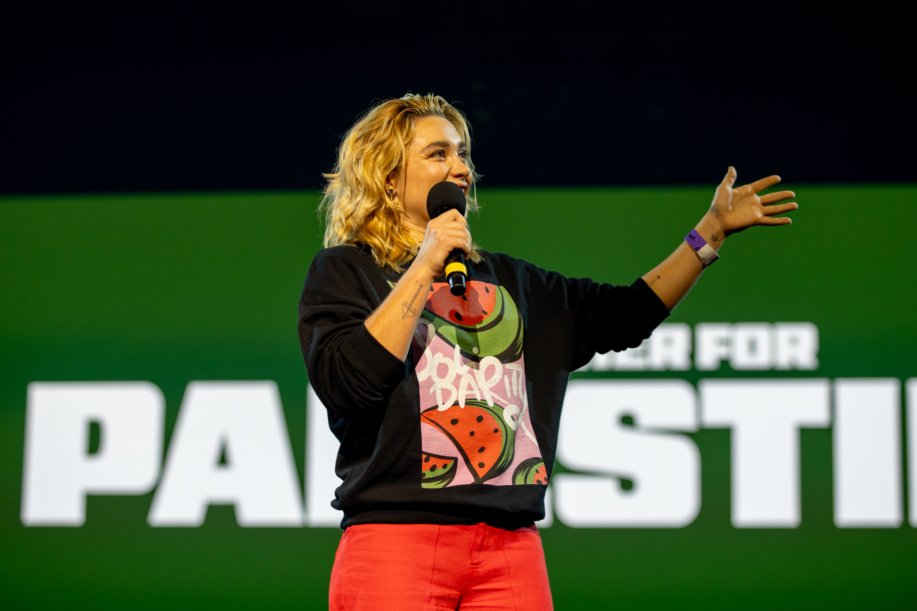 Woman with blonde curly hair holding microphone, wearing black jacket over graphic t-shirt and red trousers, speaking on stage with green backdrop and white "PALESTI" text visible.