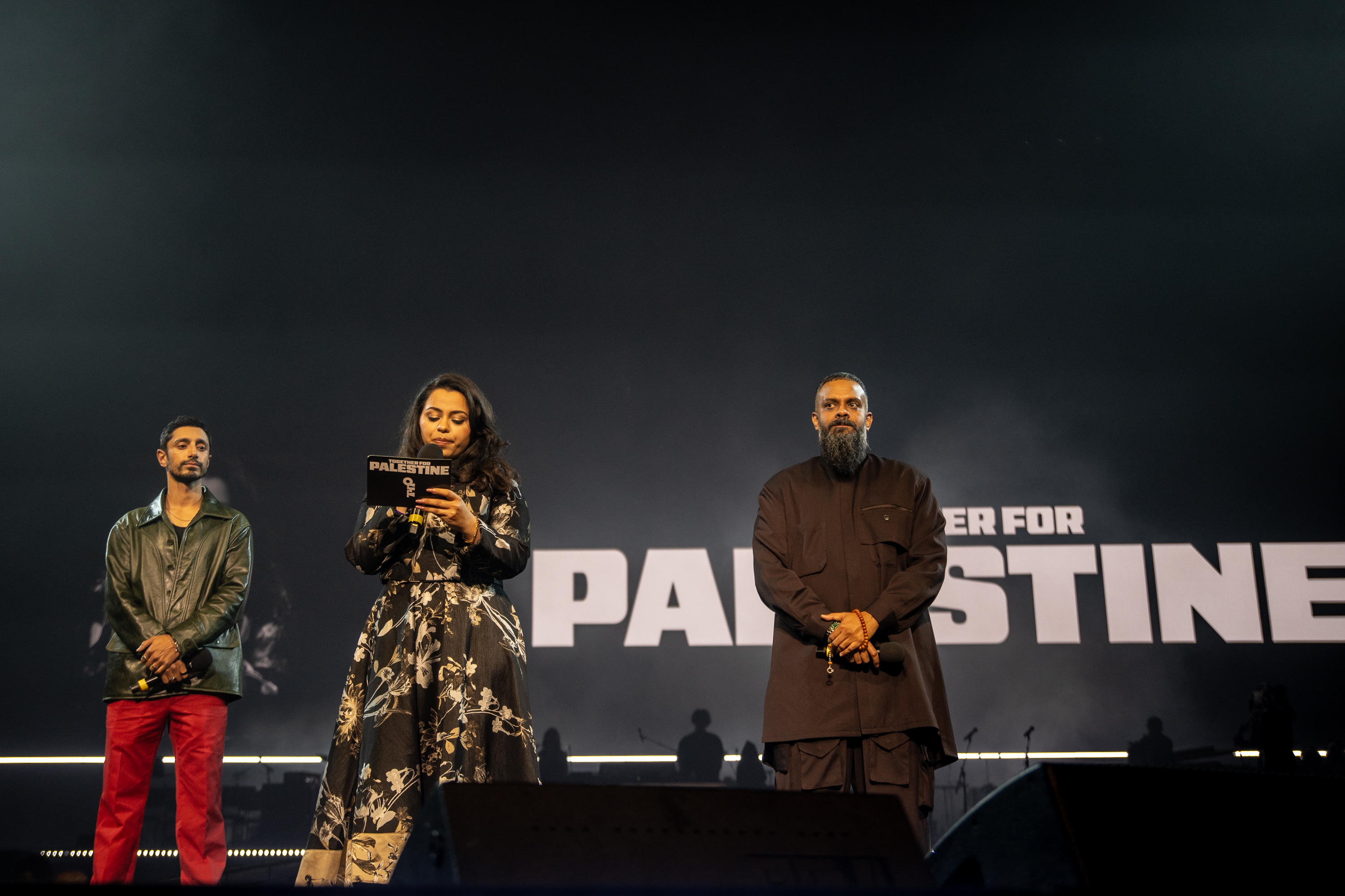 Three people on stage with "PALESTINE" text visible on backdrop. Left: man in green jacket and red trousers. Centre: woman in patterned dress holding microphone. Right: bearded man in dark clothing.