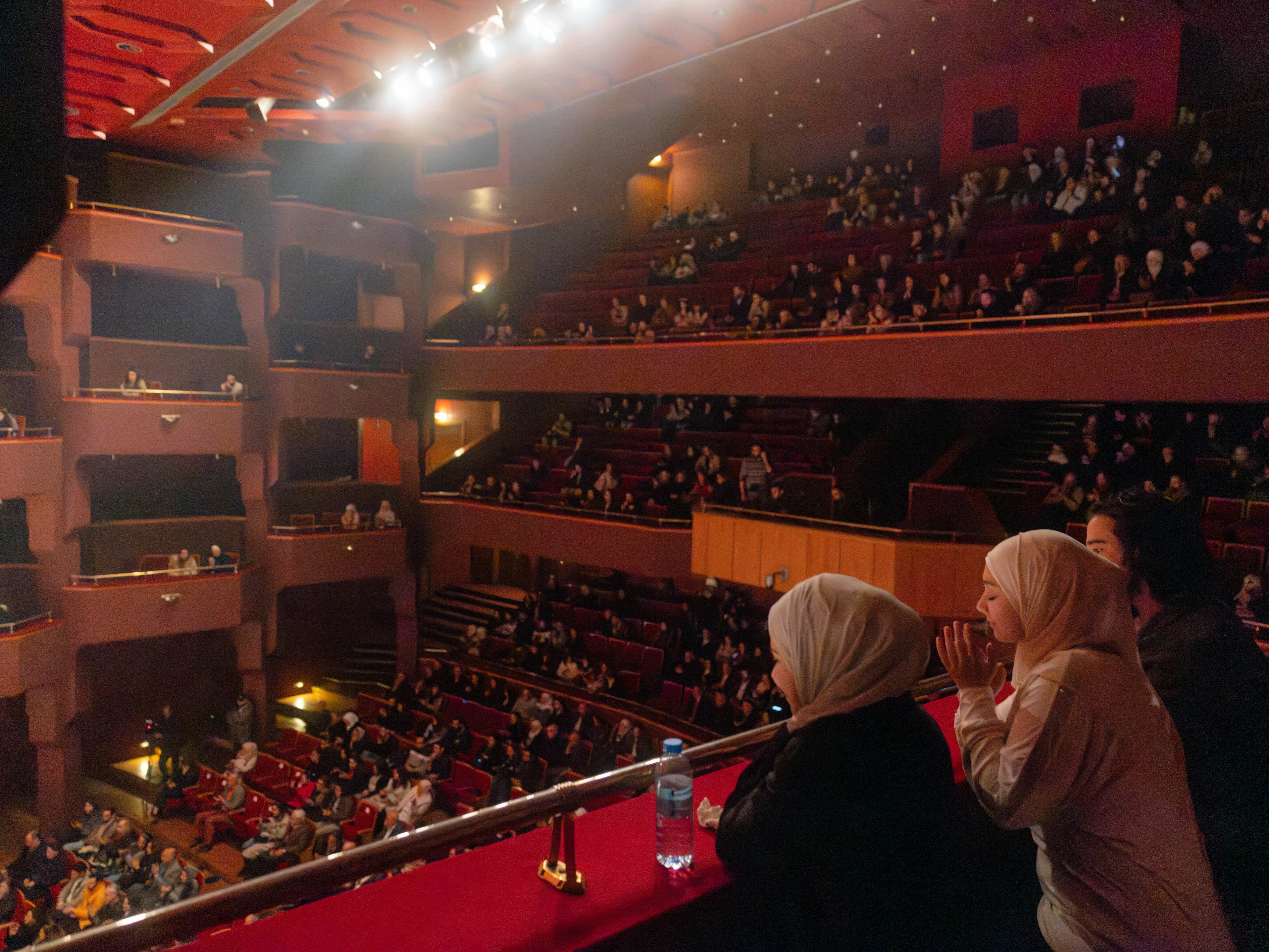 Crowded theatre auditorium with red and ornate decor, filled with seated audience members and two women standing on the balcony.