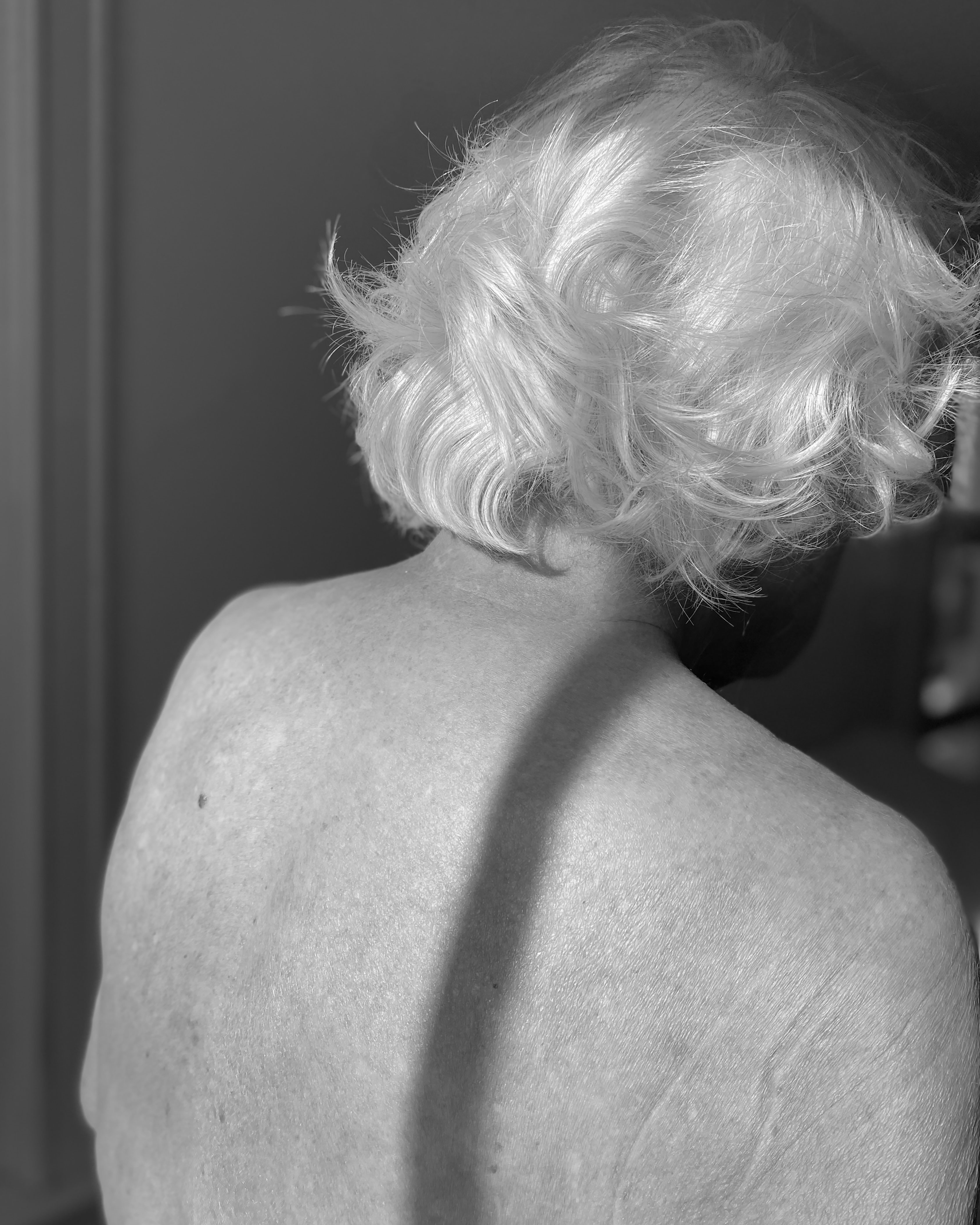 Black and white image showing the back view of a person with short, curly blonde hair and bare shoulders.