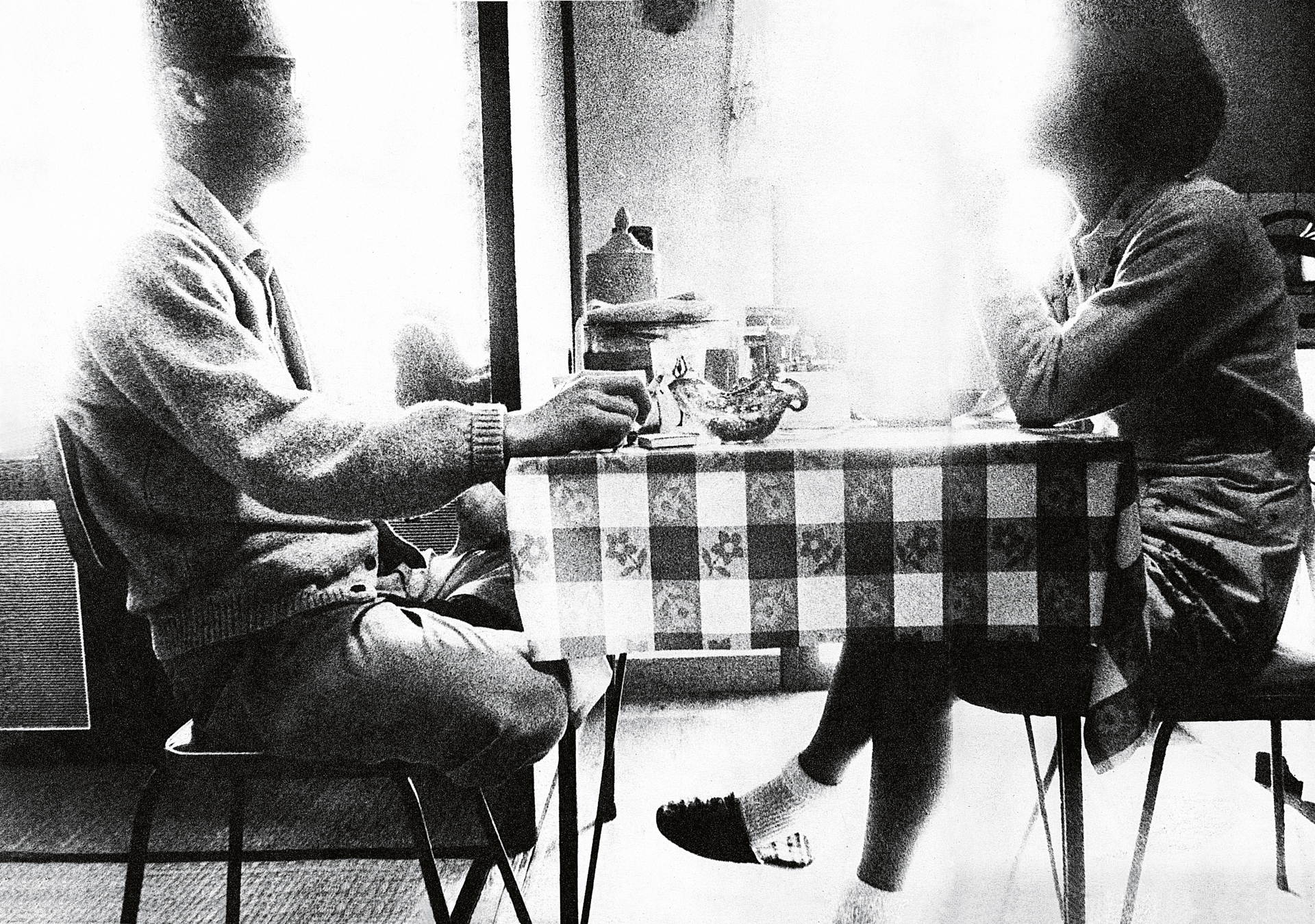 Two people seated at small table with chequered tablecloth, drinks visible, black and white photograph with soft lighting from window.
