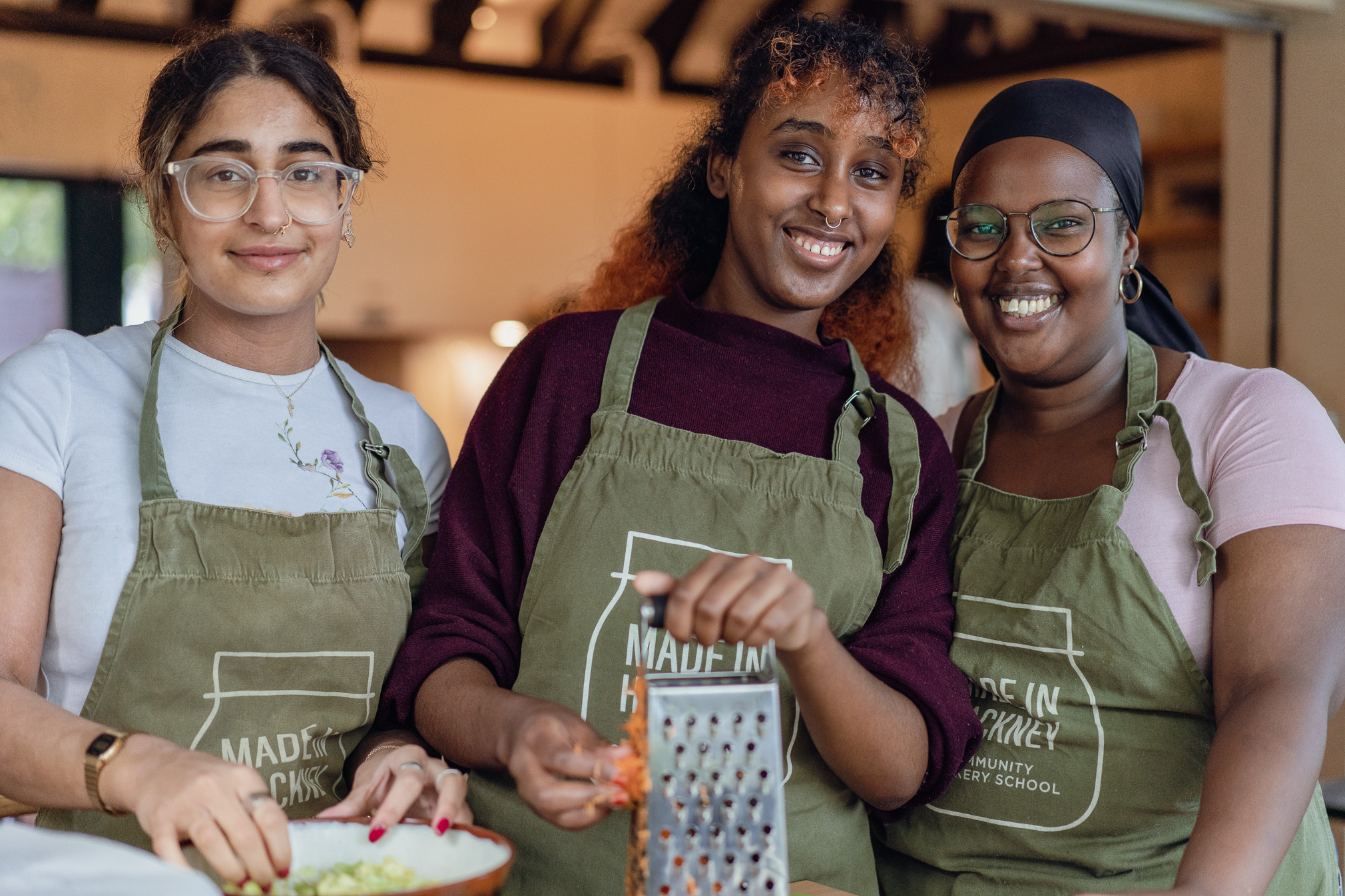 Three women wearing matching green aprons smile at camera in kitchen, one holding cheese grater, warm lighting in background.