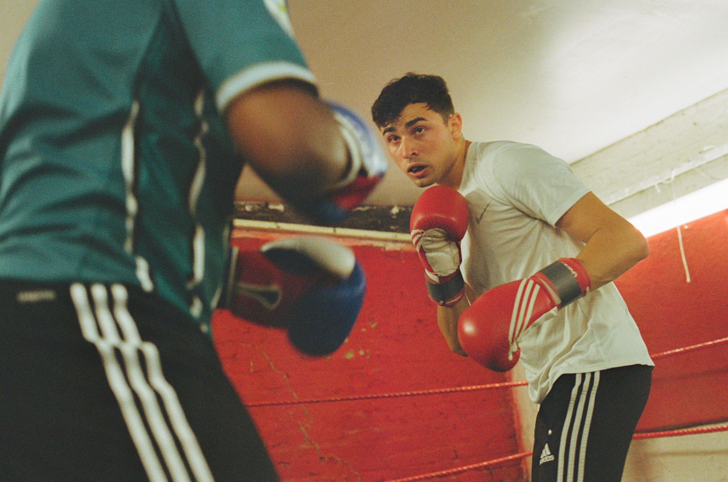 Two boxers sparring in ring with red ropes. One in green shirt throws punch whilst other in white shirt with red gloves defends.