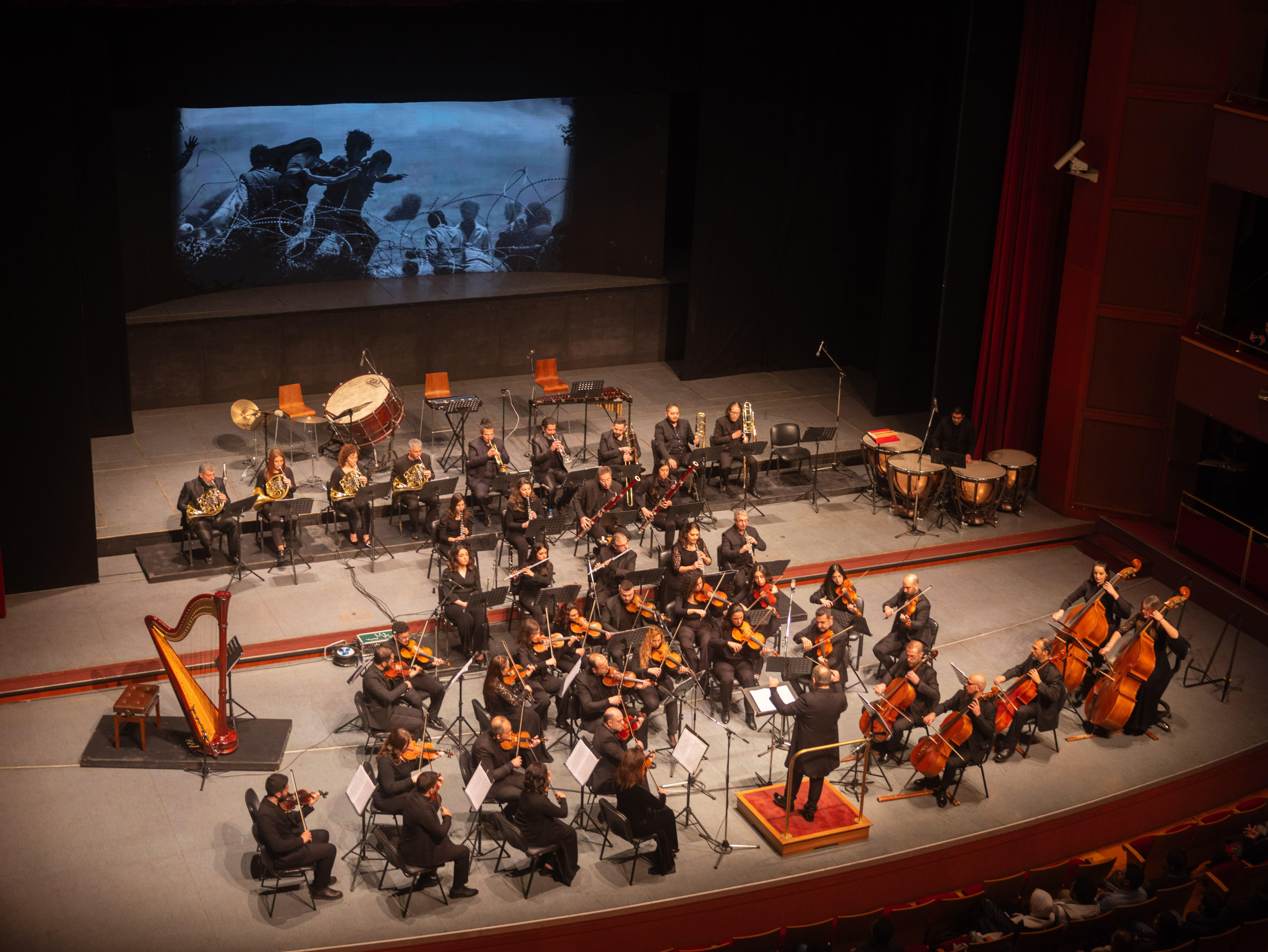 Large orchestra performing on a stage, with various musical instruments and a conductor in the centre.