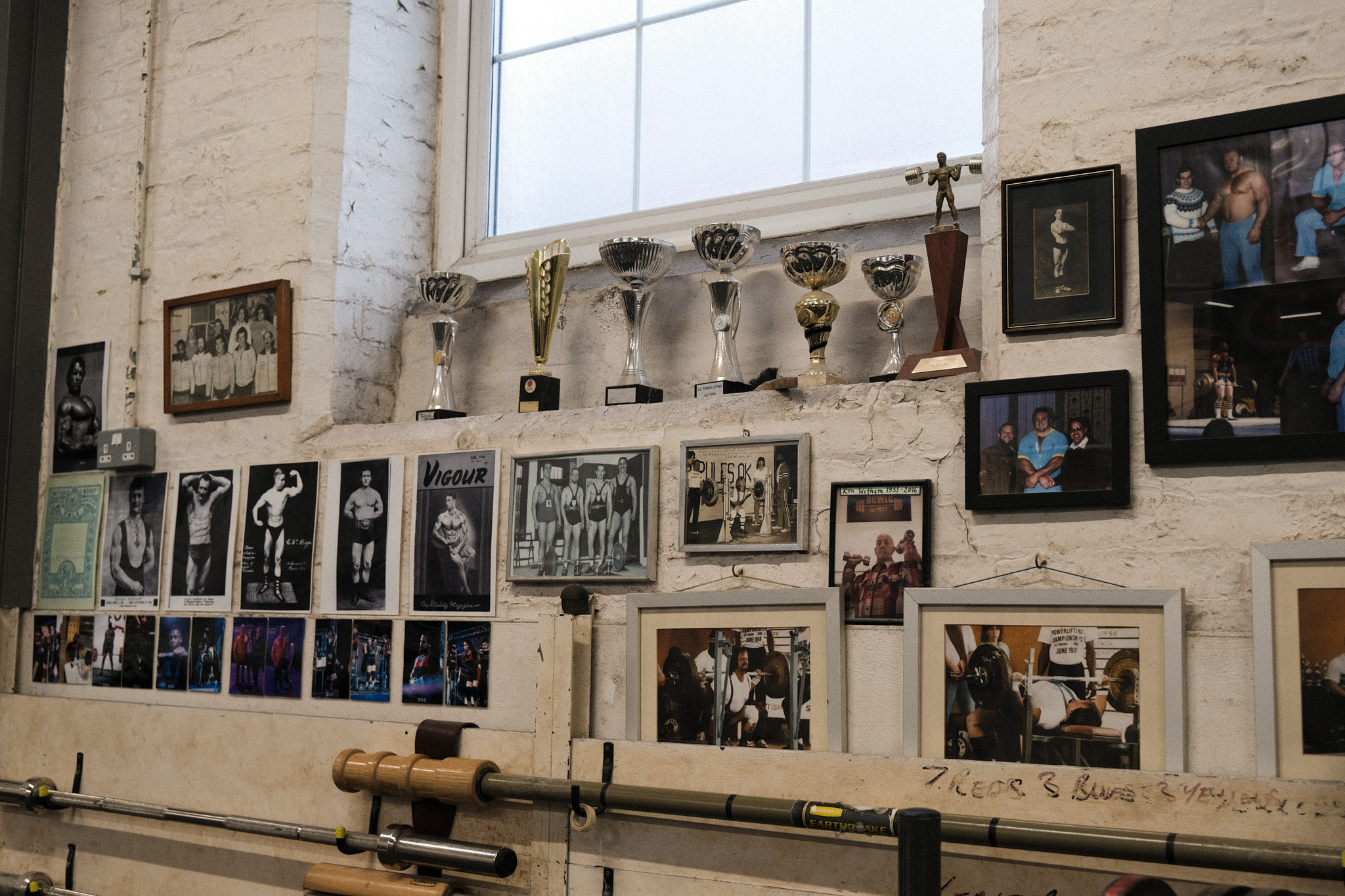 Interior wall displaying multiple framed photographs and several silver trophies on a shelf, with exposed brick and a large window above.