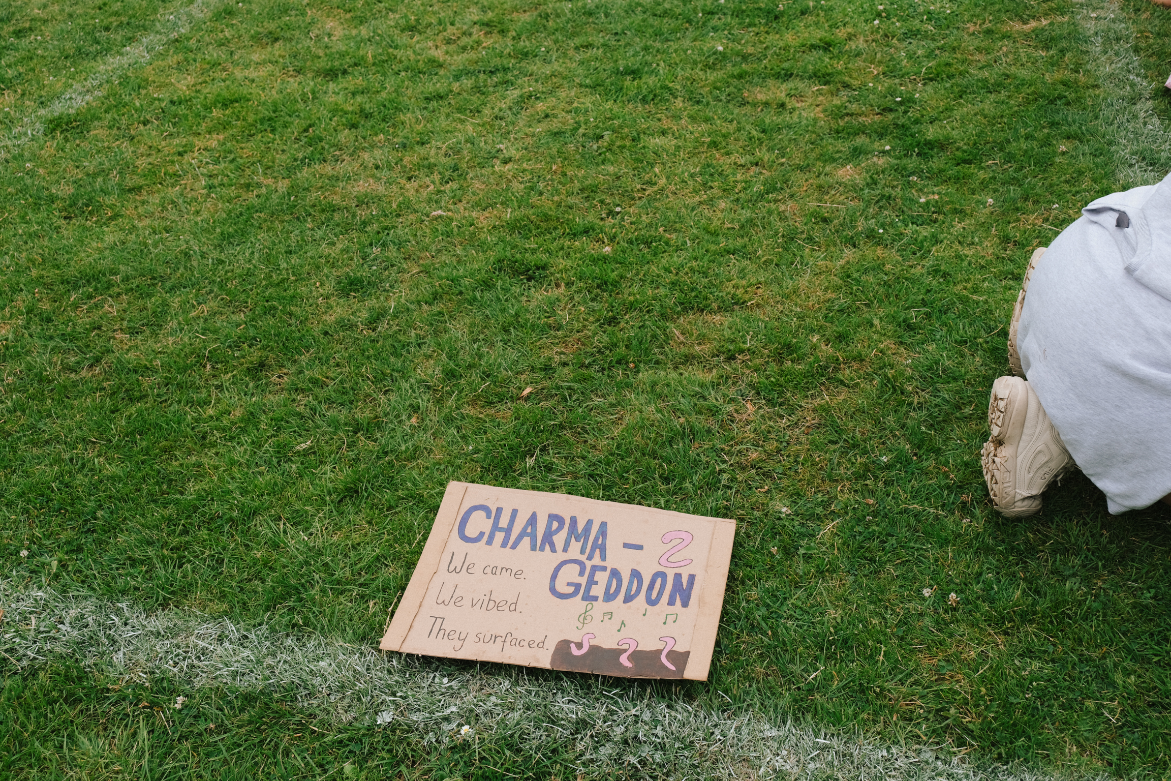 Cardboard sign reading "CHARMAGEDDON" lies on green grass next to person in white clothing kneeling on ground.