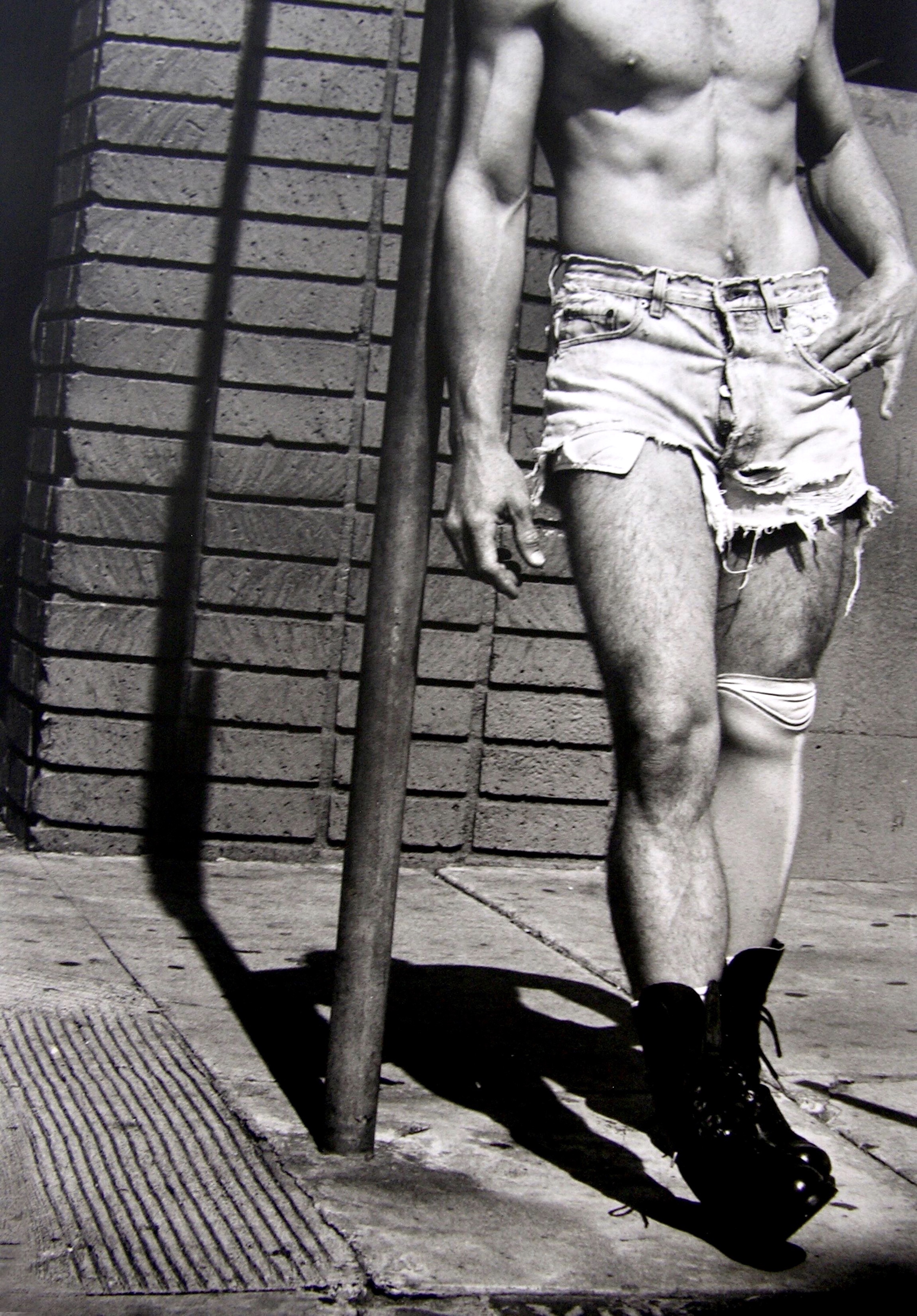 Shirtless man in distressed denim shorts and black boots leaning against metal railings on concrete steps.